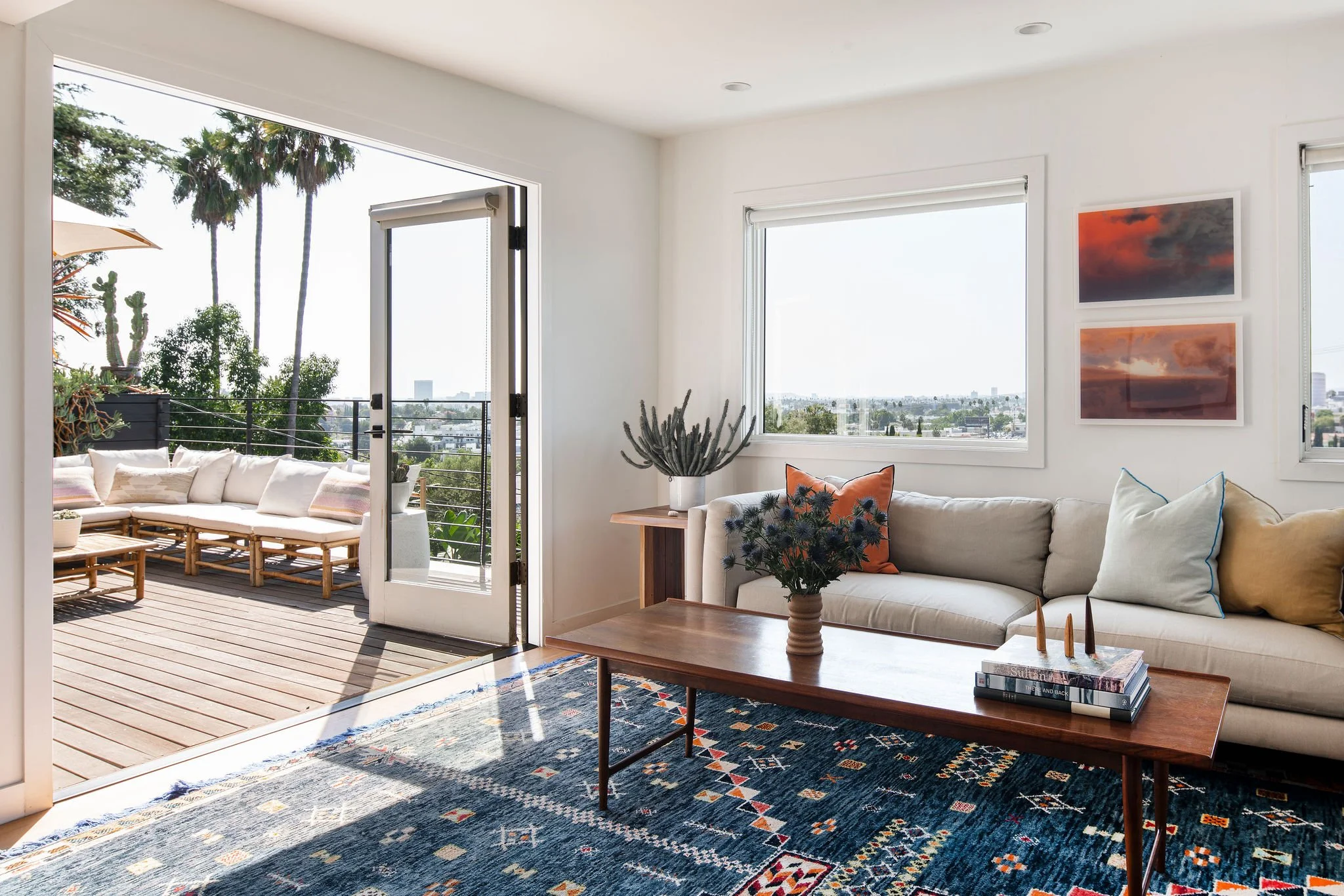 Living room with white sofa, colorful pillows, wooden coffee table with books and candles, patterned rug, potted plants, large windows showing cityscape, and a balcony with outdoor seating and palm trees.