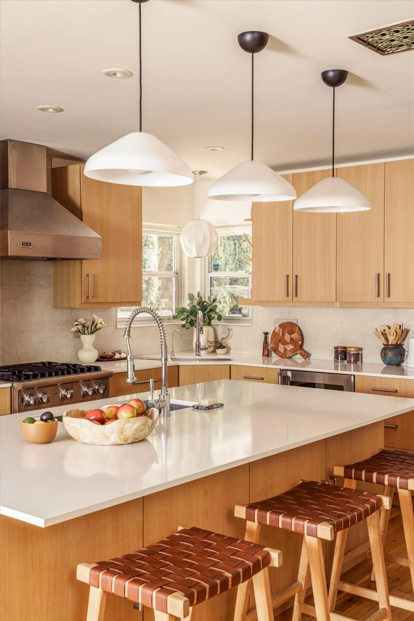 A modern kitchen with wooden cabinets, white countertops, and three hanging pendant lights. There are windows behind the sink with plants and kitchen utensils, and a bowl of apples on the island counter with three wooden barstools.