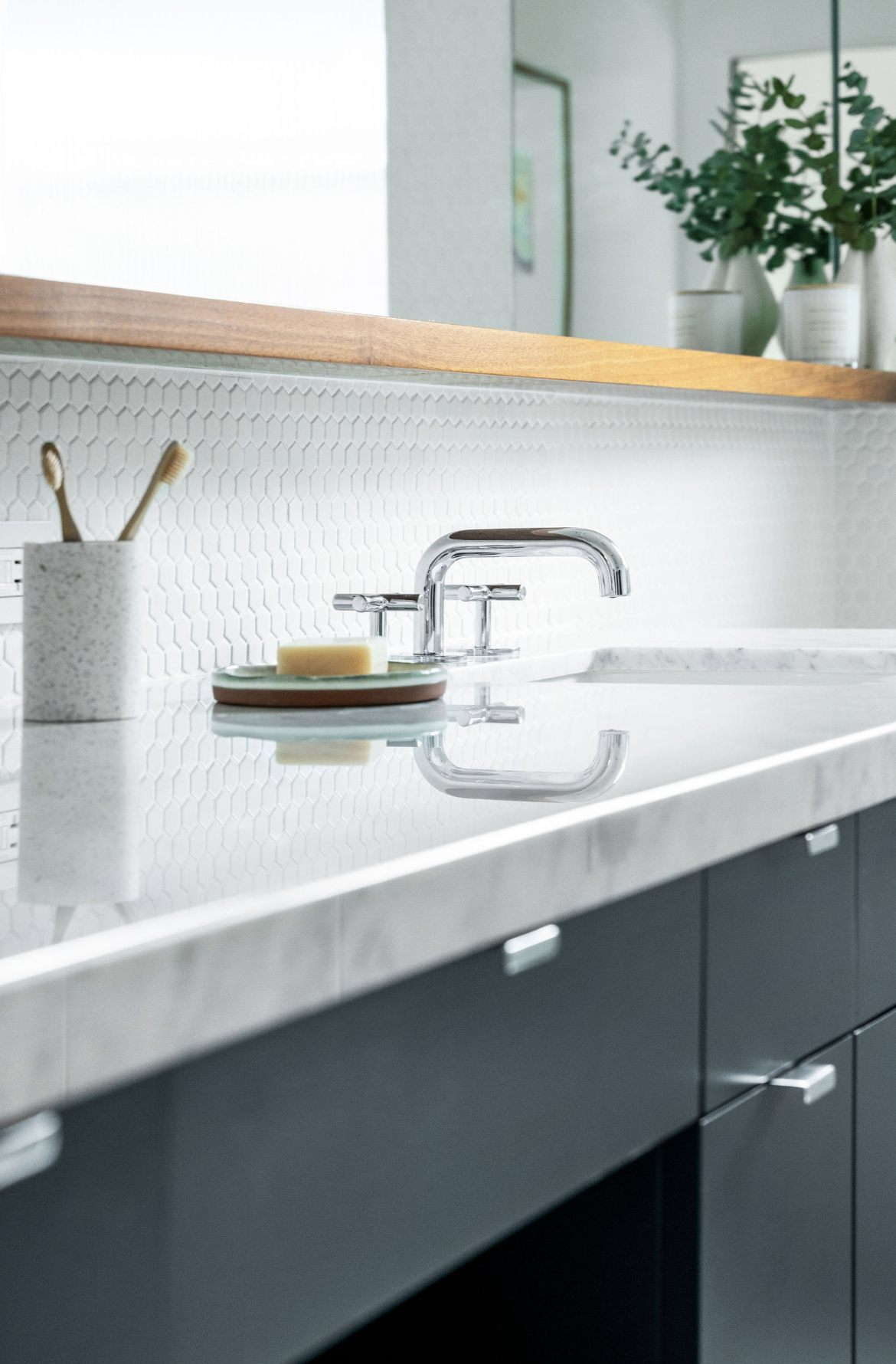 Modern kitchen with white hexagonal tile backsplash, white marble countertop, and black cabinets. Small soap and a toothbrush holder are near the faucet, and there are green plants in white pots in the background.