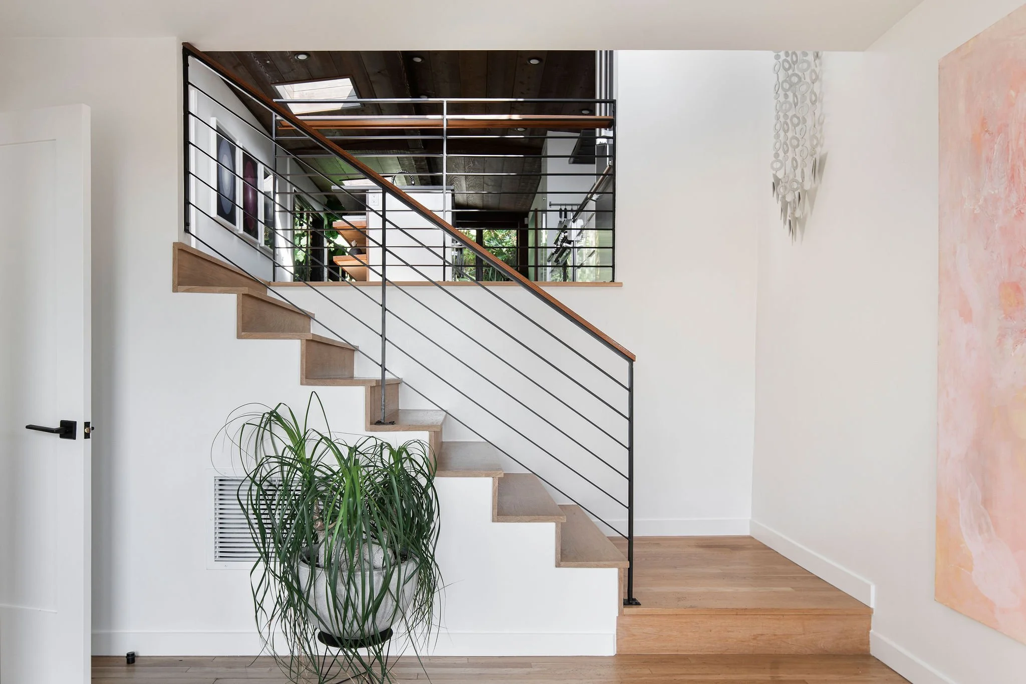 Interior view of a modern staircase with wooden steps, black metal railing, a potted green plant, white walls, and artwork.