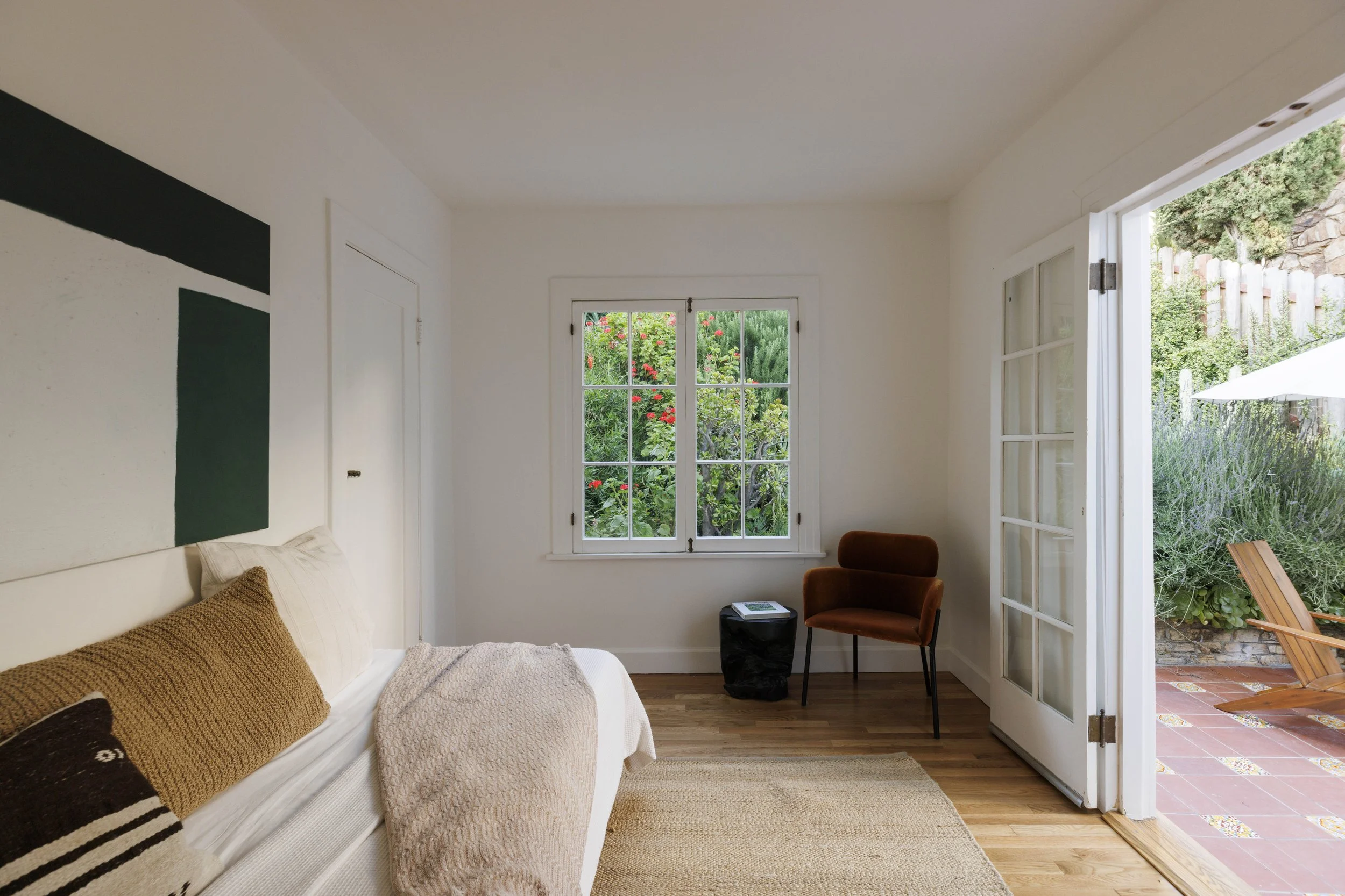 Cozy bedroom with white walls, a window showing greenery outside, a bed with beige and brown pillows, and a patio door leading to an outdoor area with a wooden chair and umbrella.