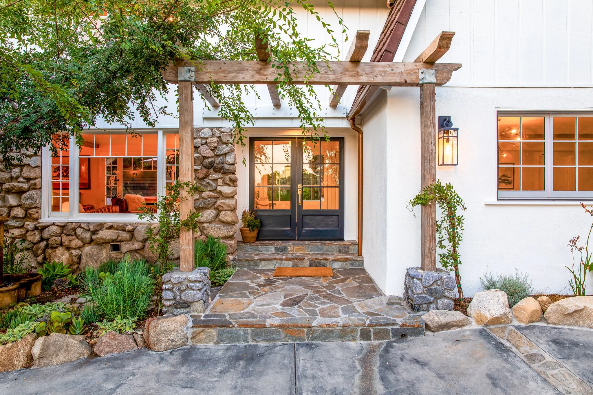 Front porch of a house with stone and white wall exterior, black double front doors, stone steps, potted plants, and decorative wooden beams overhead.
