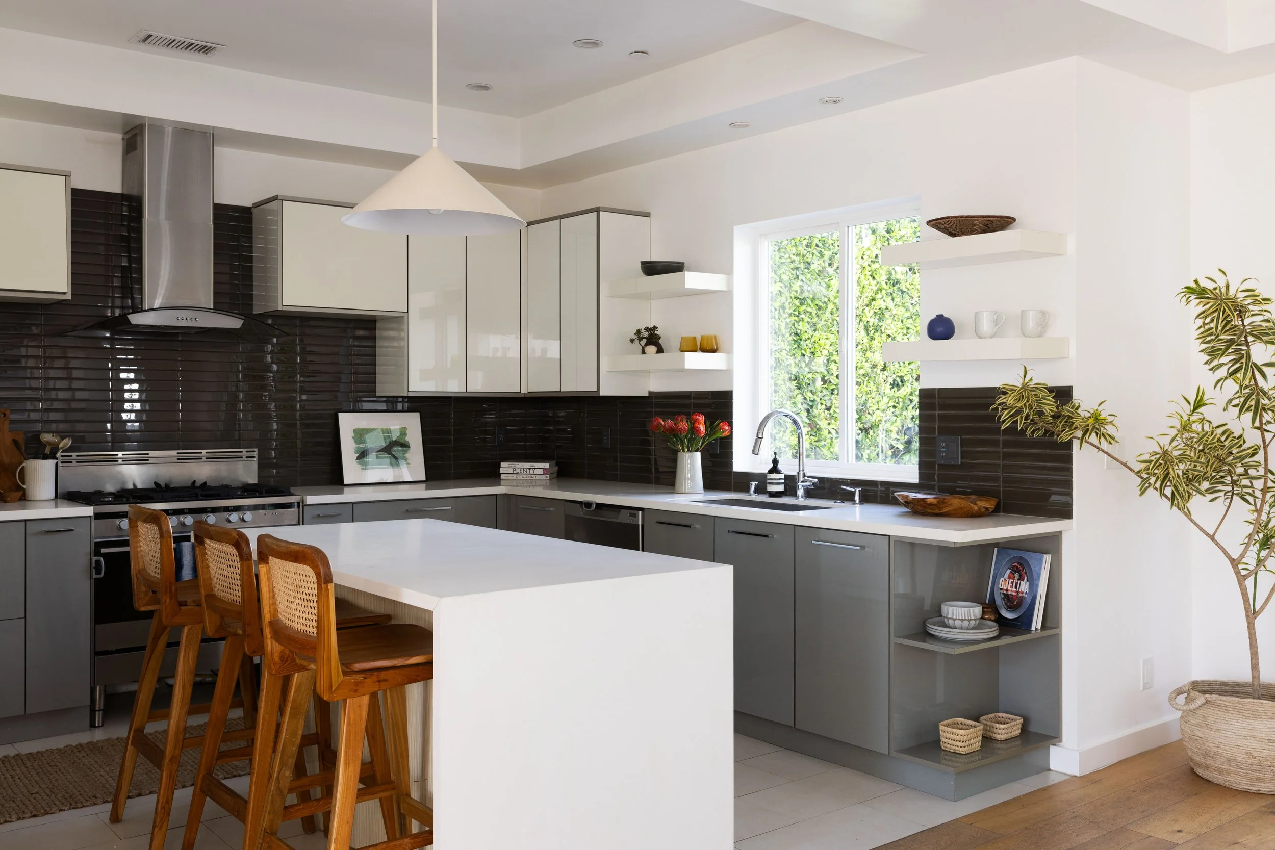 Modern kitchen with gray and white cabinets, black backsplash, wooden chairs, and a white island. Decor includes flowers, bowls, and shelves with vases, with a large window letting in natural light.