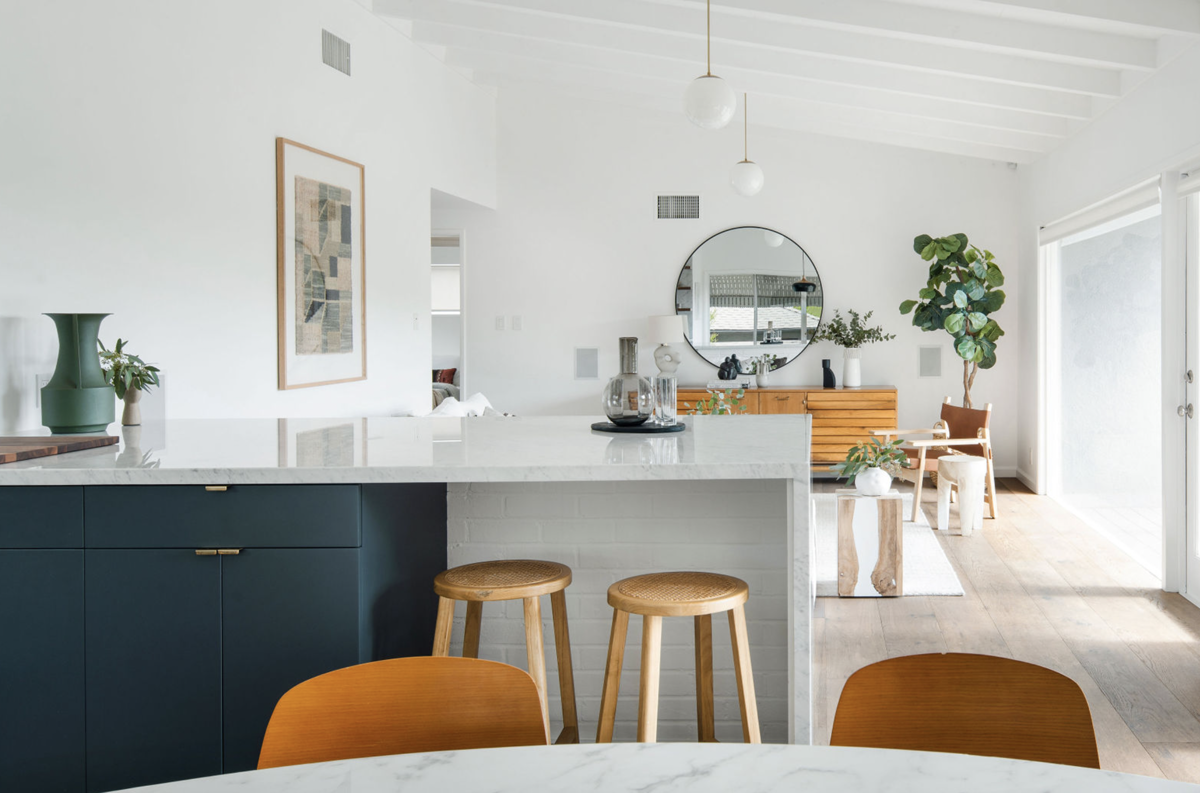 Open-concept kitchen and dining area with white walls, wooden flooring, modern decor, and large sliding glass door to patio, featuring a kitchen island with dark cabinets, wooden stools, and a round mirror on the wall.