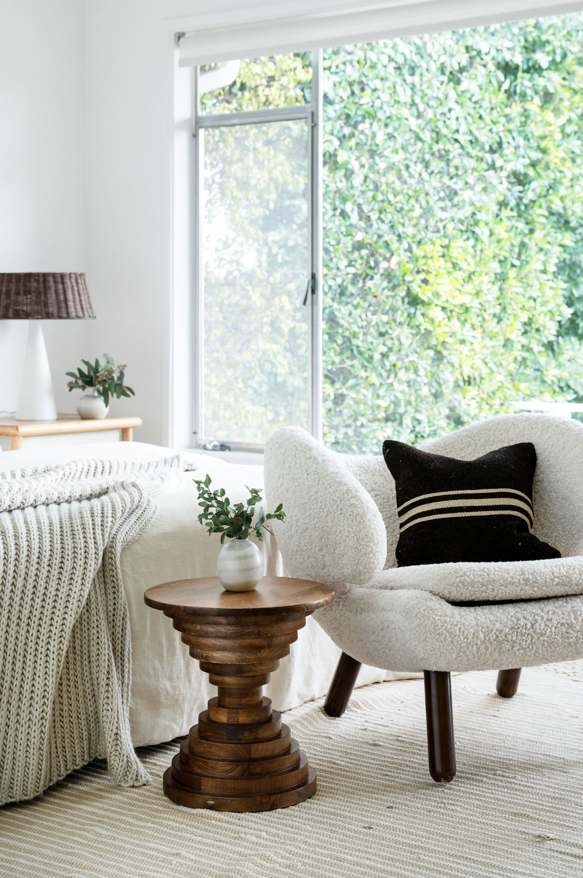 Bright living room featuring a white armchair with a black-striped pillow, a wooden side table with a small vase of greenery, a bed with a chunky knit blanket, a bedside table with a lamp, and large window showing green trees outside.