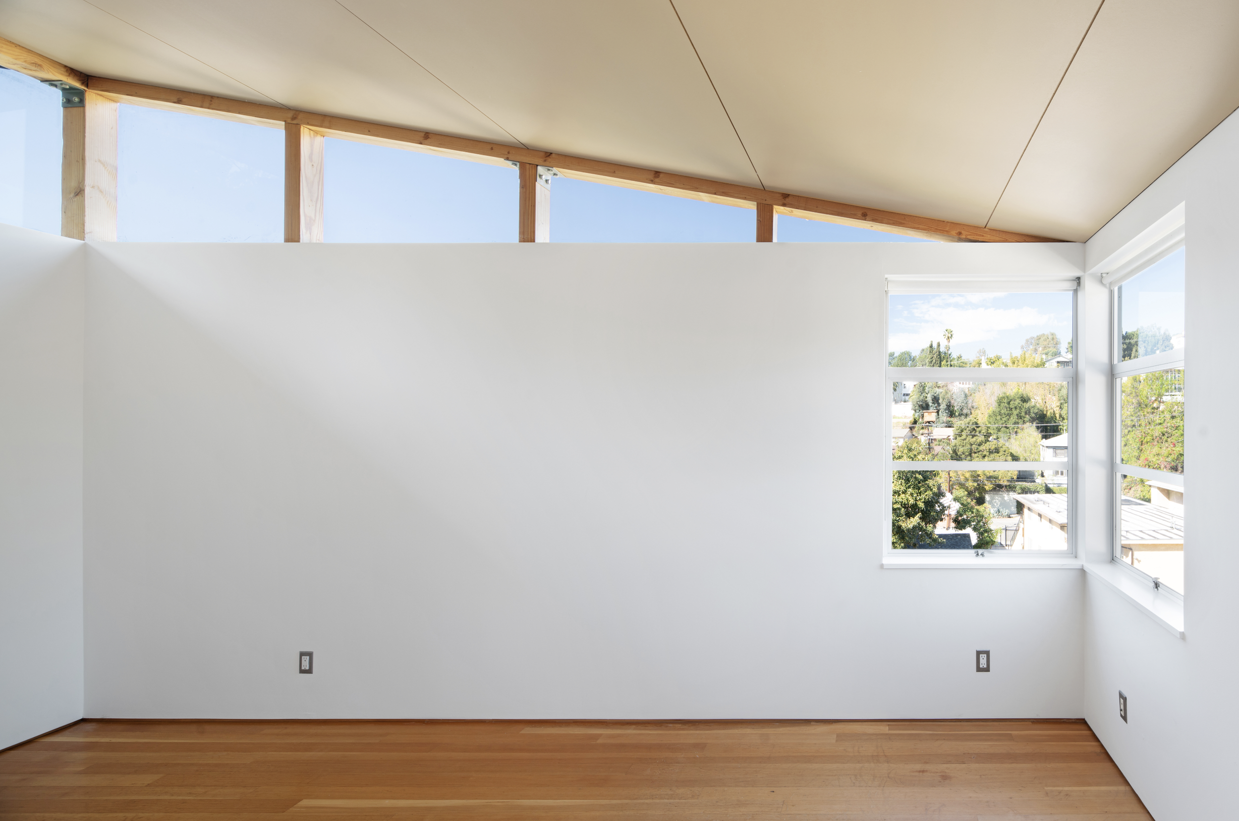 Empty room with white walls, wooden floor, large windows showing an outdoor view, and an unfinished ceiling with exposed wooden beams.