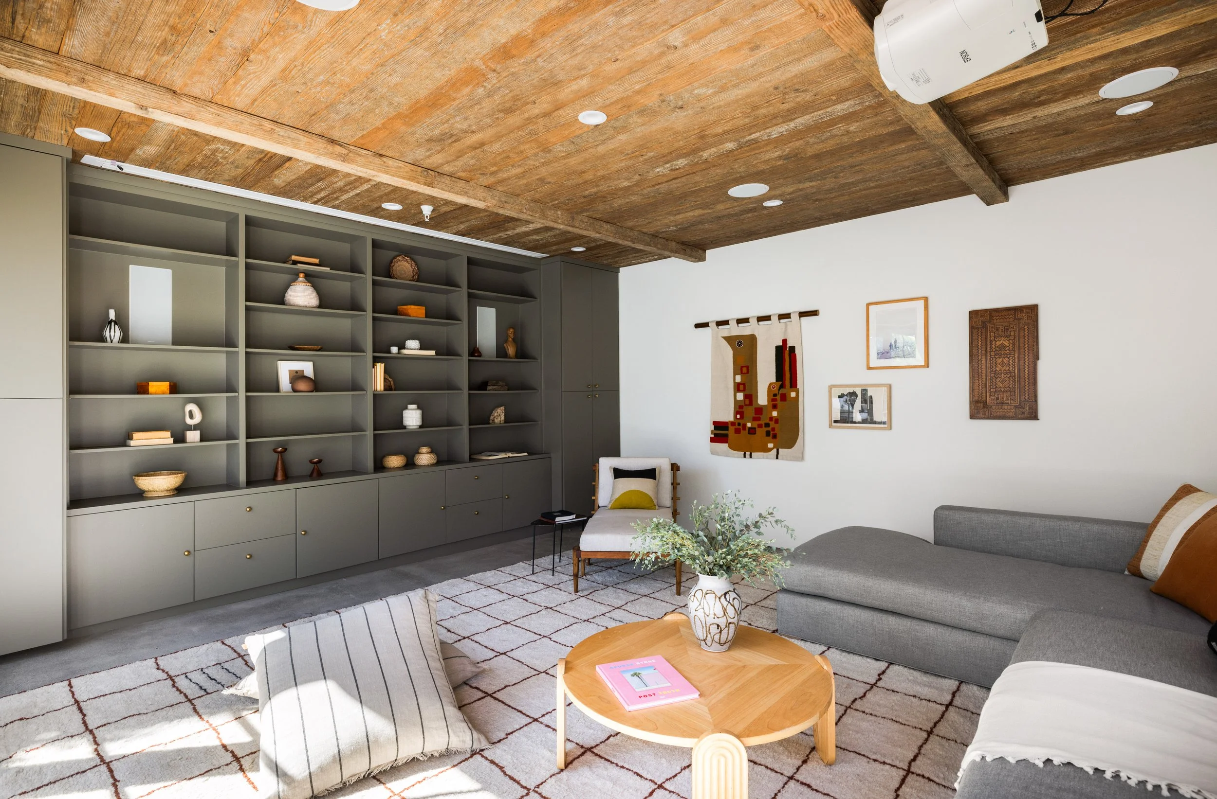 Modern living room with gray built-in shelving unit, white walls, wooden ceiling, gray sectional sofa with pillows, round wooden coffee table with a pink book and a decorative vase, armchair, patterned rug, and wall art including a woven piece and a 