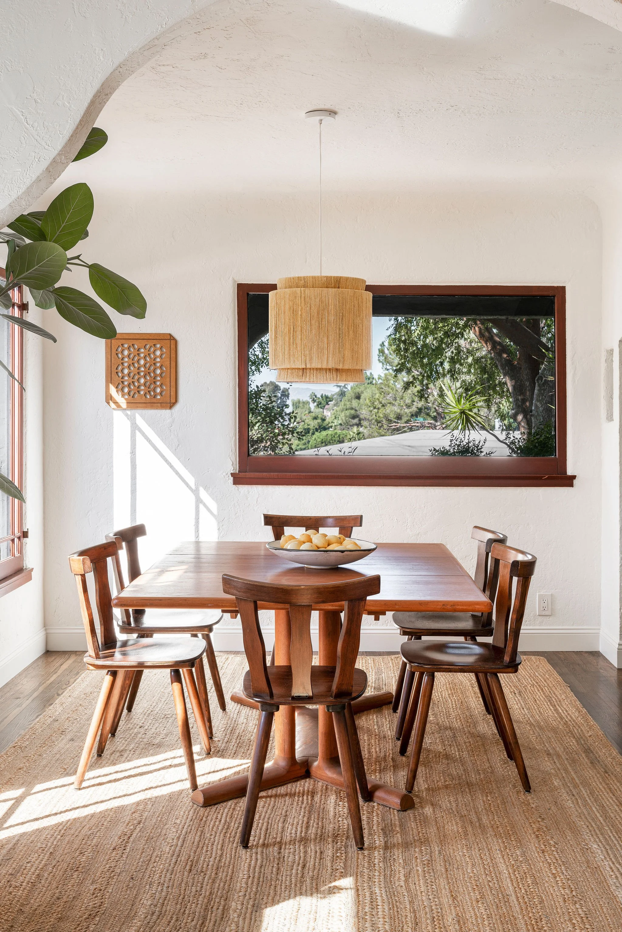 A dining room with a wooden table and five matching chairs. There is a bowl of fruit on the table, a large window showing greenery outside, and natural sunlight illuminating the space.