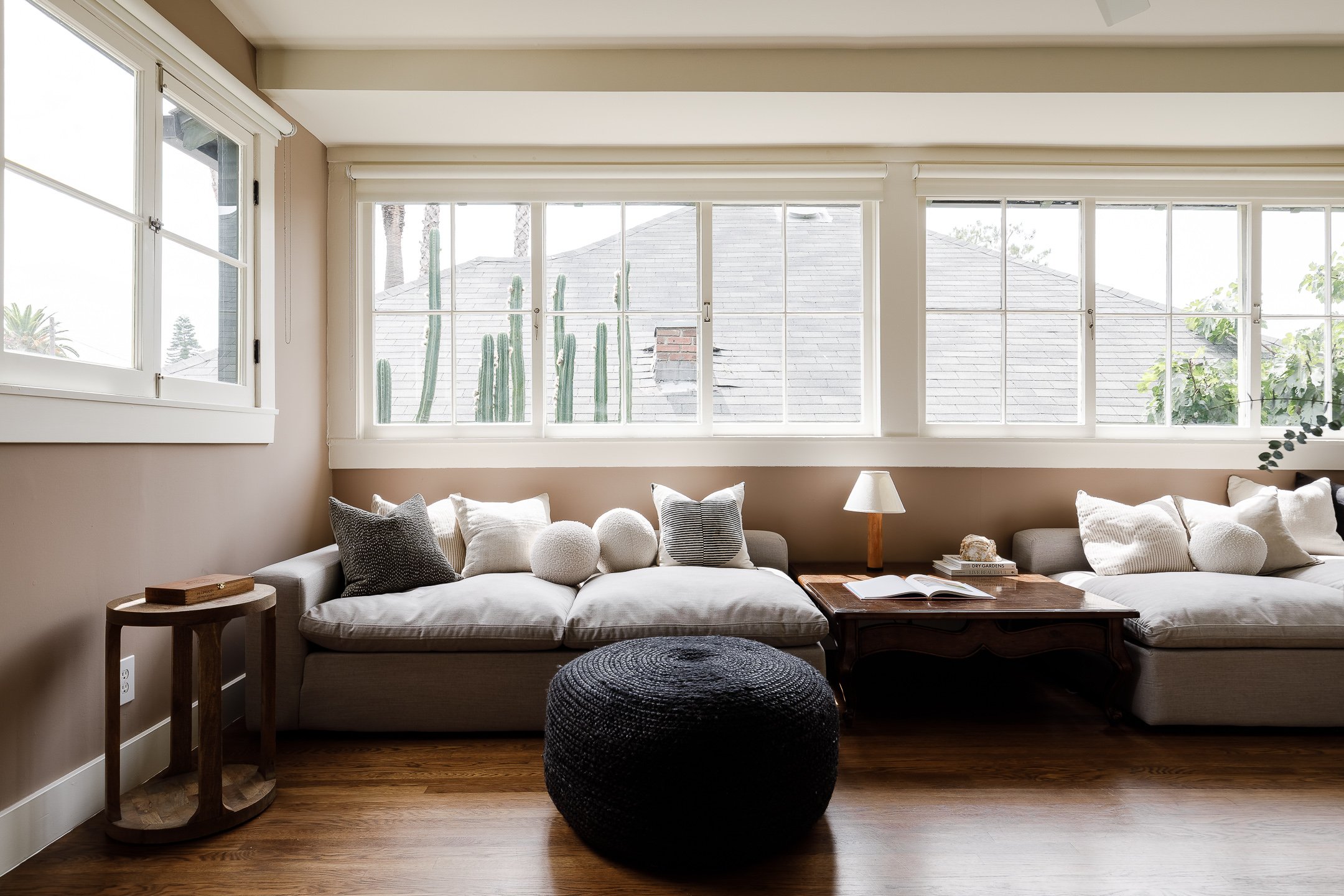 Bright living room with large windows, beige walls, gray sofa with various pillows, round black pouf, wooden side table, and a small table with a lamp and open book.