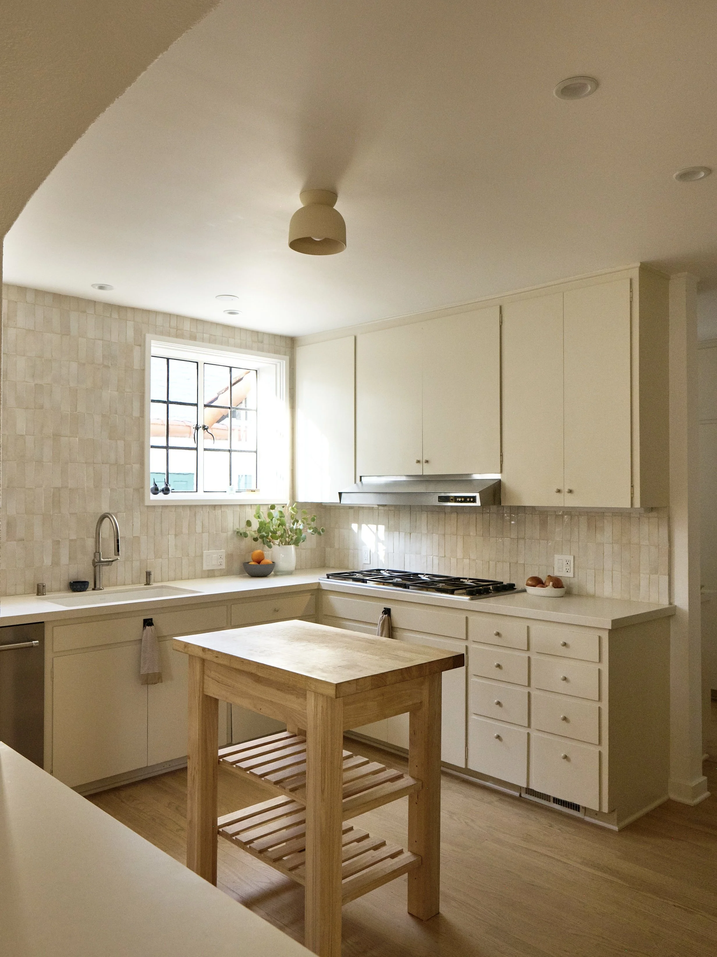 A kitchen with off-white cabinets, a small wooden island, a window above the sink, a stovetop, and some potted plants and bowls on the counter.