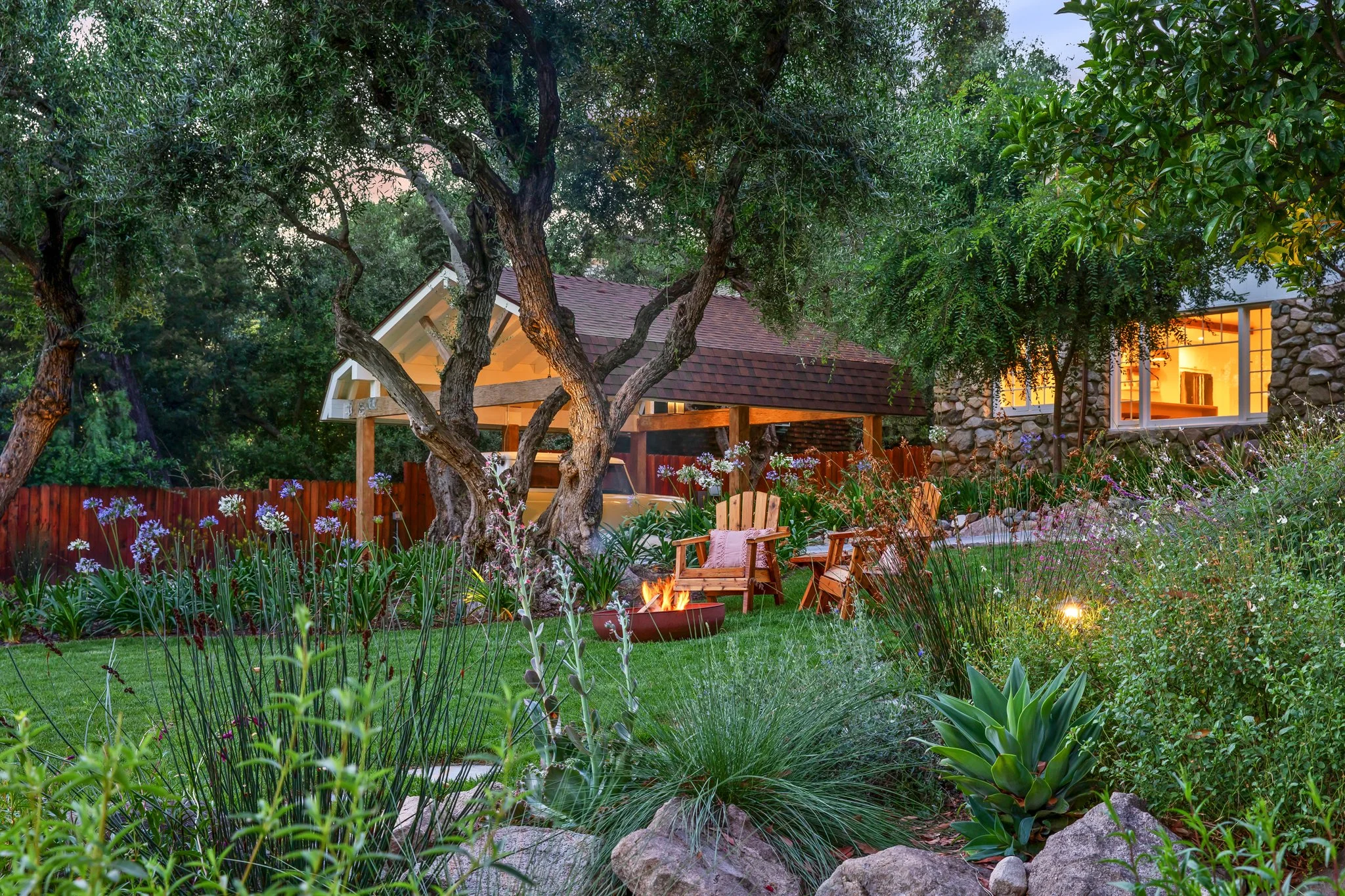 A backyard garden with lush greenery, trees, flowering plants, a fire pit with chairs, and a house with a large window illuminated from inside.