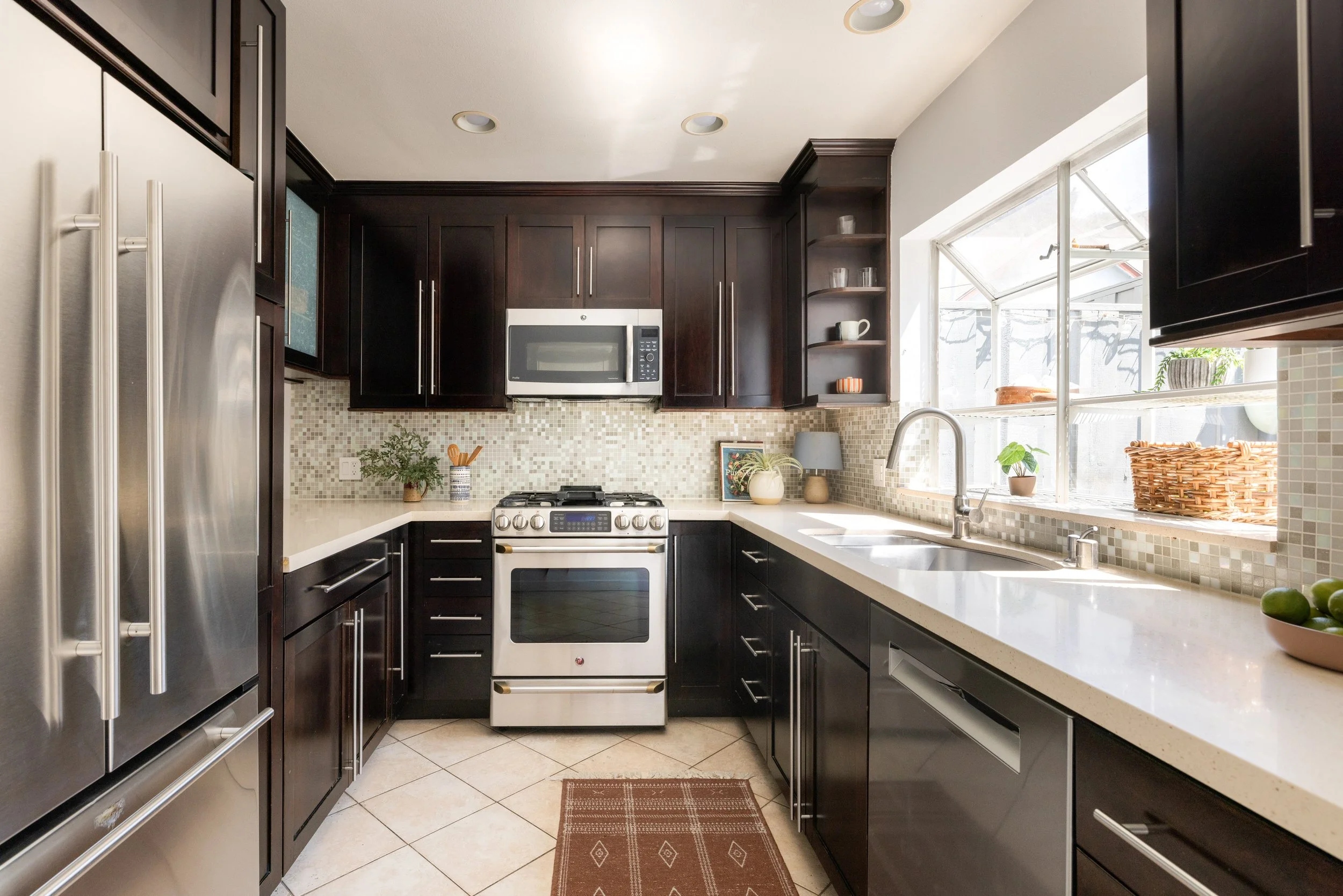 Modern kitchen with dark wood cabinets, stainless steel refrigerator, microwave, and stove. White countertops, mosaic tile backsplash, and a large window above the sink with plants and wicker baskets.