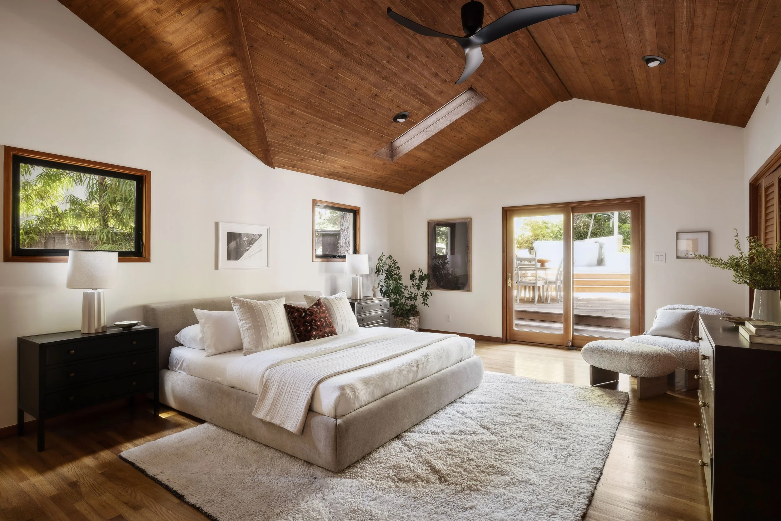 A spacious bedroom with a high wooden ceiling, a large bed with white linens and pillows, two black nightstands with lamps, sliding glass doors leading to a deck, framed windows, and minimal decor.