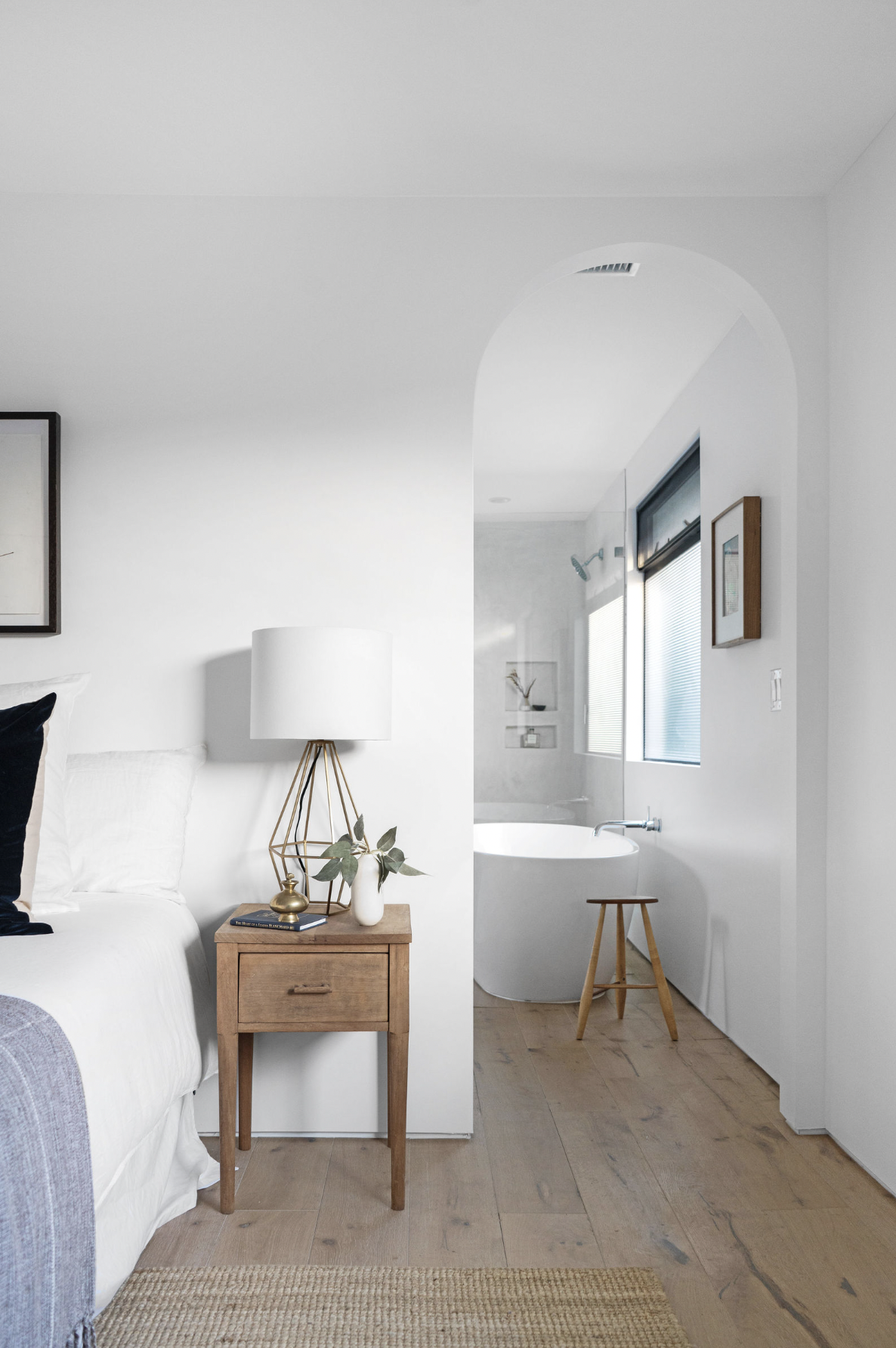 A cozy bedroom with a nightstand, lamp, and decorative items, connected to a bathroom with a bathtub and natural light.
