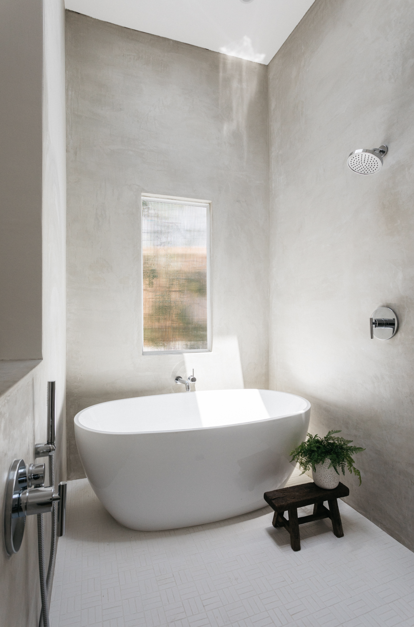 Modern bathroom with a freestanding white bathtub, a small wooden stool with a plant, a frosted window, and a rain shower head on the wall.