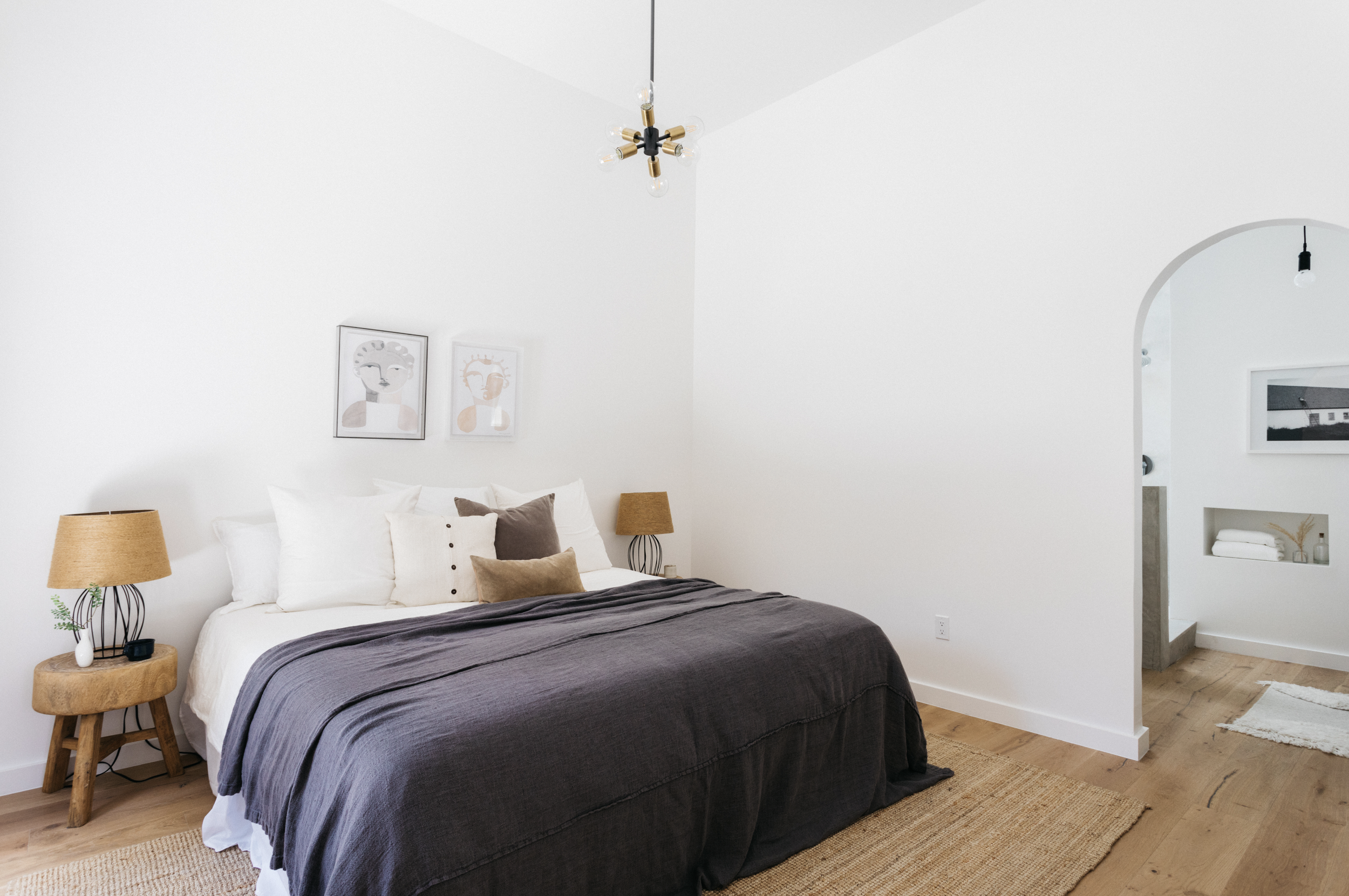 A bedroom with white walls and a bed with white and gray bedding, plus multiple pillows. Two bedside tables with lamps are on either side of the bed. Framed artwork hangs above the bed. An arched doorway leads to another room with a framed picture an