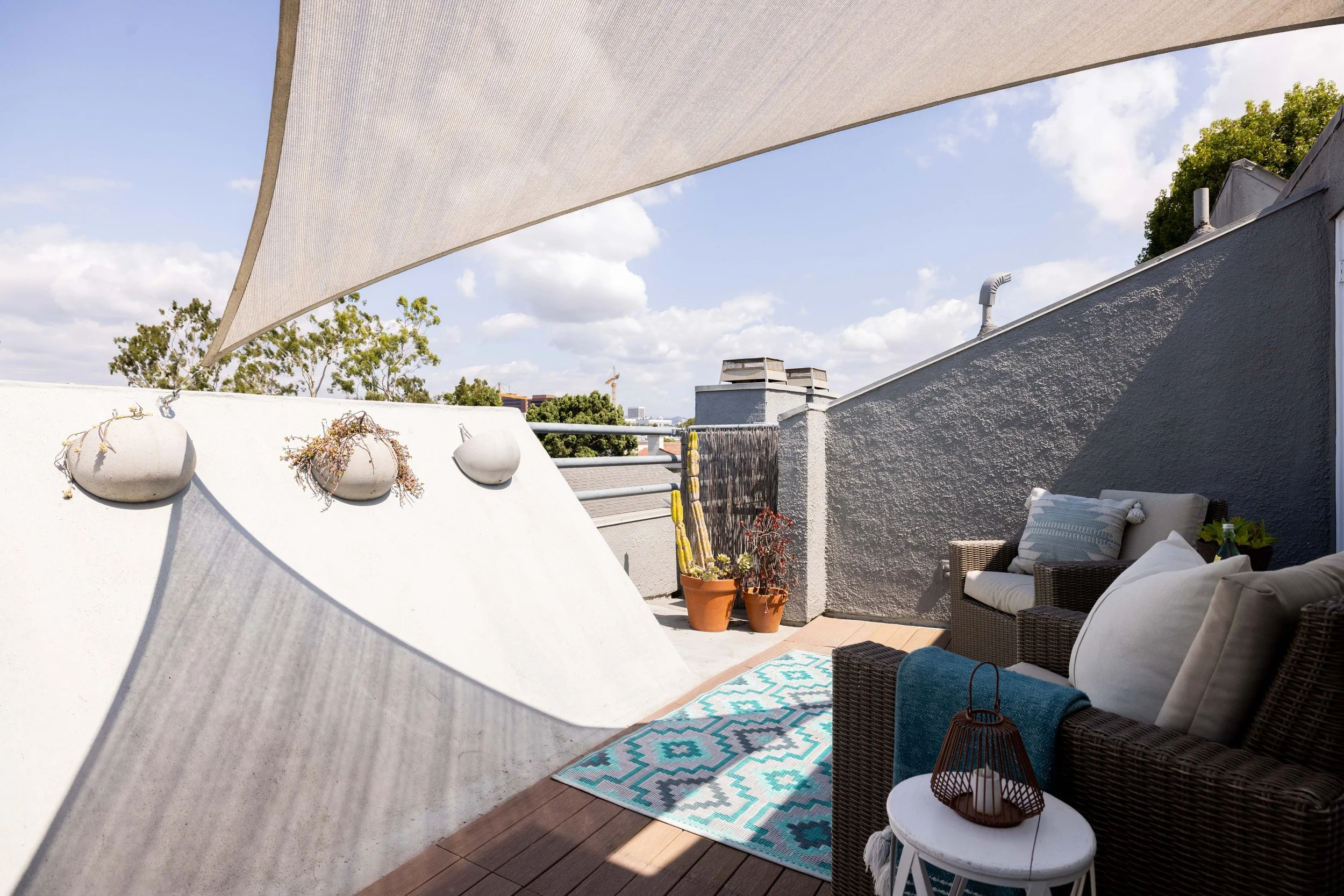 A decorated outdoor balcony with a wicker sofa, pillows, a small round table with a lantern, potted cacti, a colorful rug, and a beige canopy overhead.