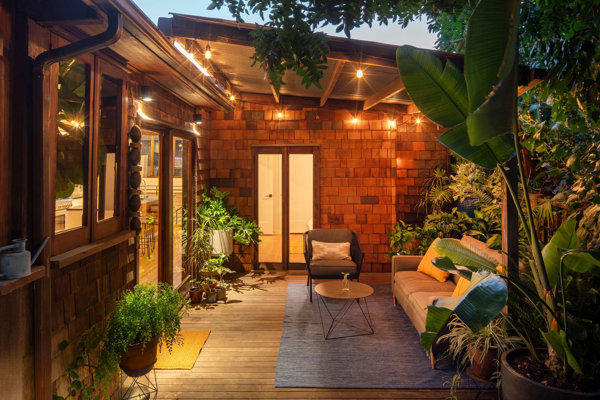 A cozy outdoor patio with wooden flooring, string lights, potted plants, a beige sofa with yellow pillows, a small round wooden table, a gray armchair with a pillow, and lush green plants surrounding the space.