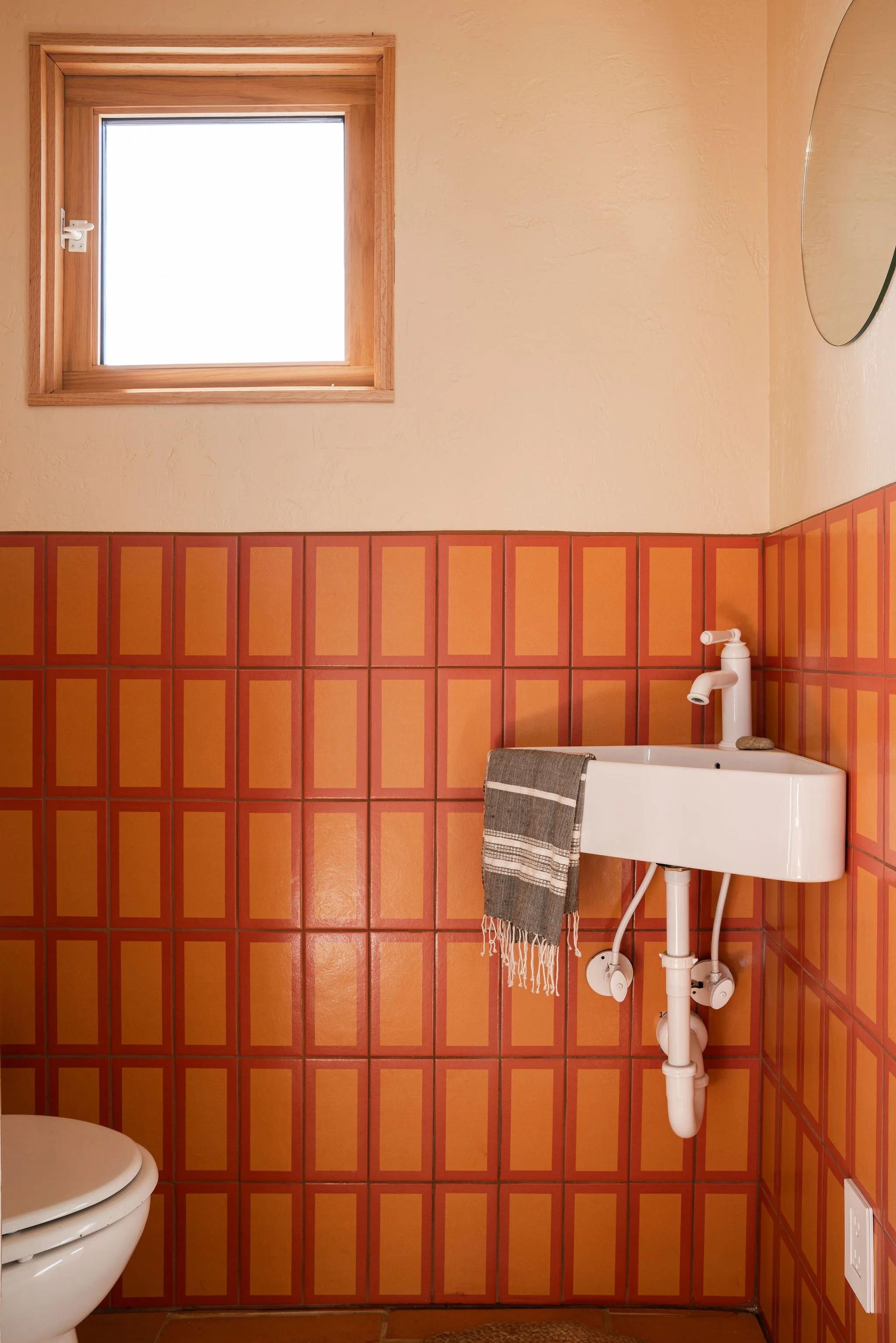 A small bathroom corner with orange and yellow patterned tiles, a white utility sink with a gray towel hanging, a small window with a wooden frame, and a portion of a white toilet visible in the lower left corner.