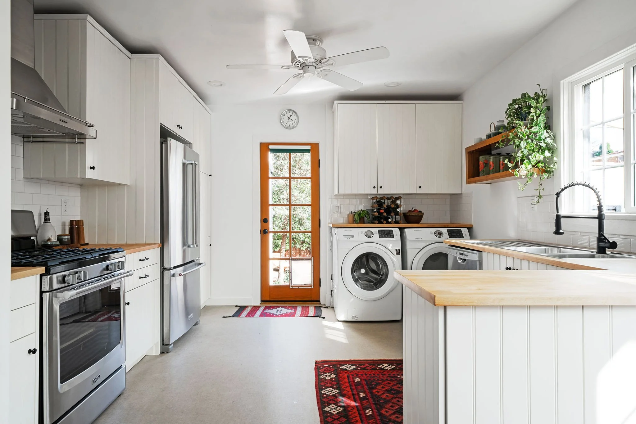Bright kitchen with white cabinets, wooden countertops, stainless steel appliances, a washer and dryer, a window above the sink, and a glass door leading outside.