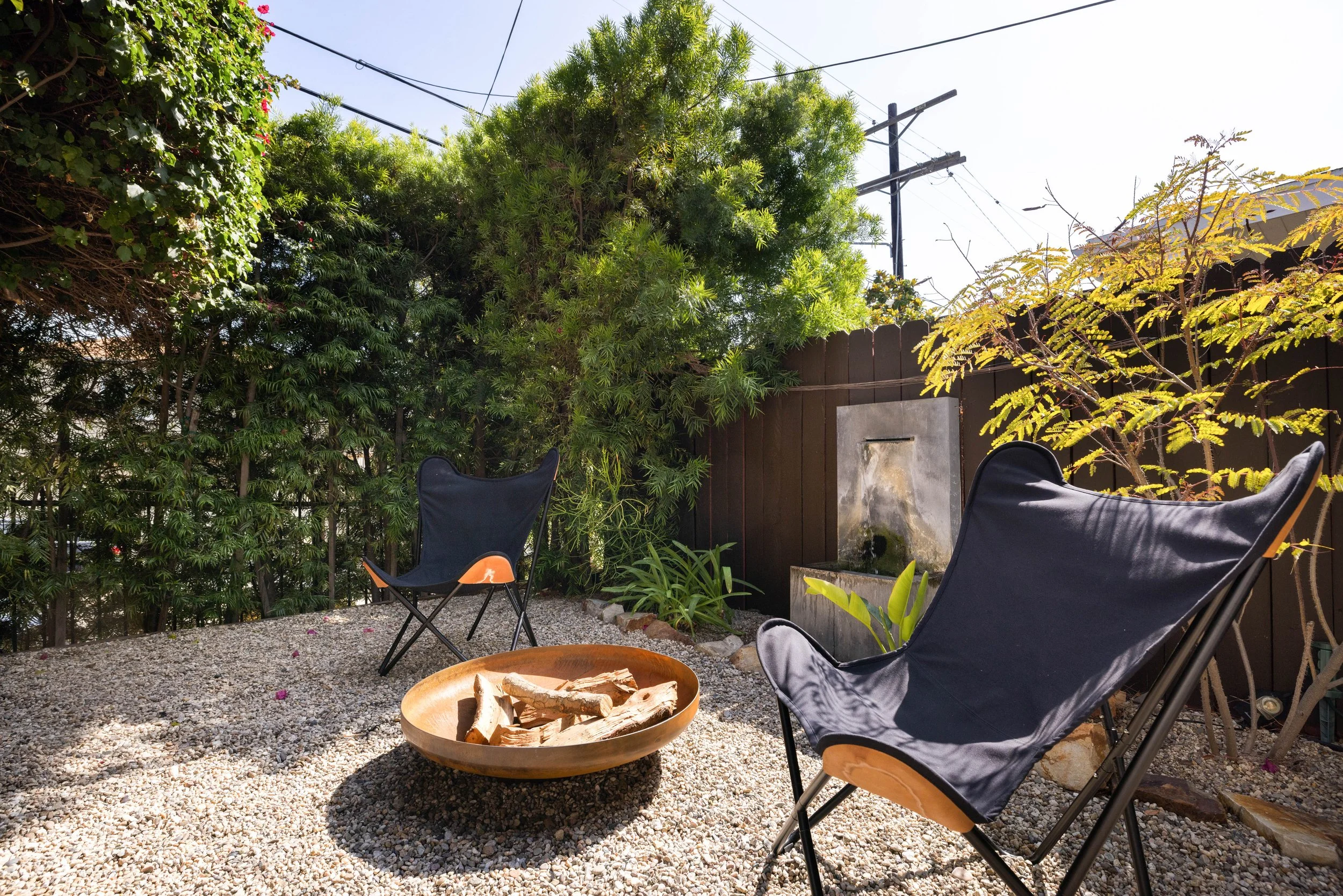 A backyard patio with two black camping chairs, a wooden fire pit with logs, greenery including trees and bushes, a wooden fence, and a water feature with water flowing down a wall, under a clear sky with overhead power lines.