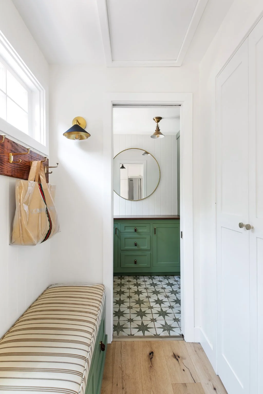 A small entryway with a striped cushioned bench, hooks with beige shopping bags, a black and brass wall sconce, a round mirror, and a doorway leading to a room with green cabinets and patterned floor tiles.