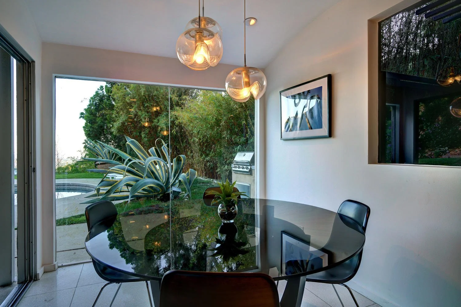 Dining room with glass table, black chairs, a small plant, and pendant lights. Large sliding glass door reveals outdoor yard with a agave plant, trees, and patio, and a grill near the door.