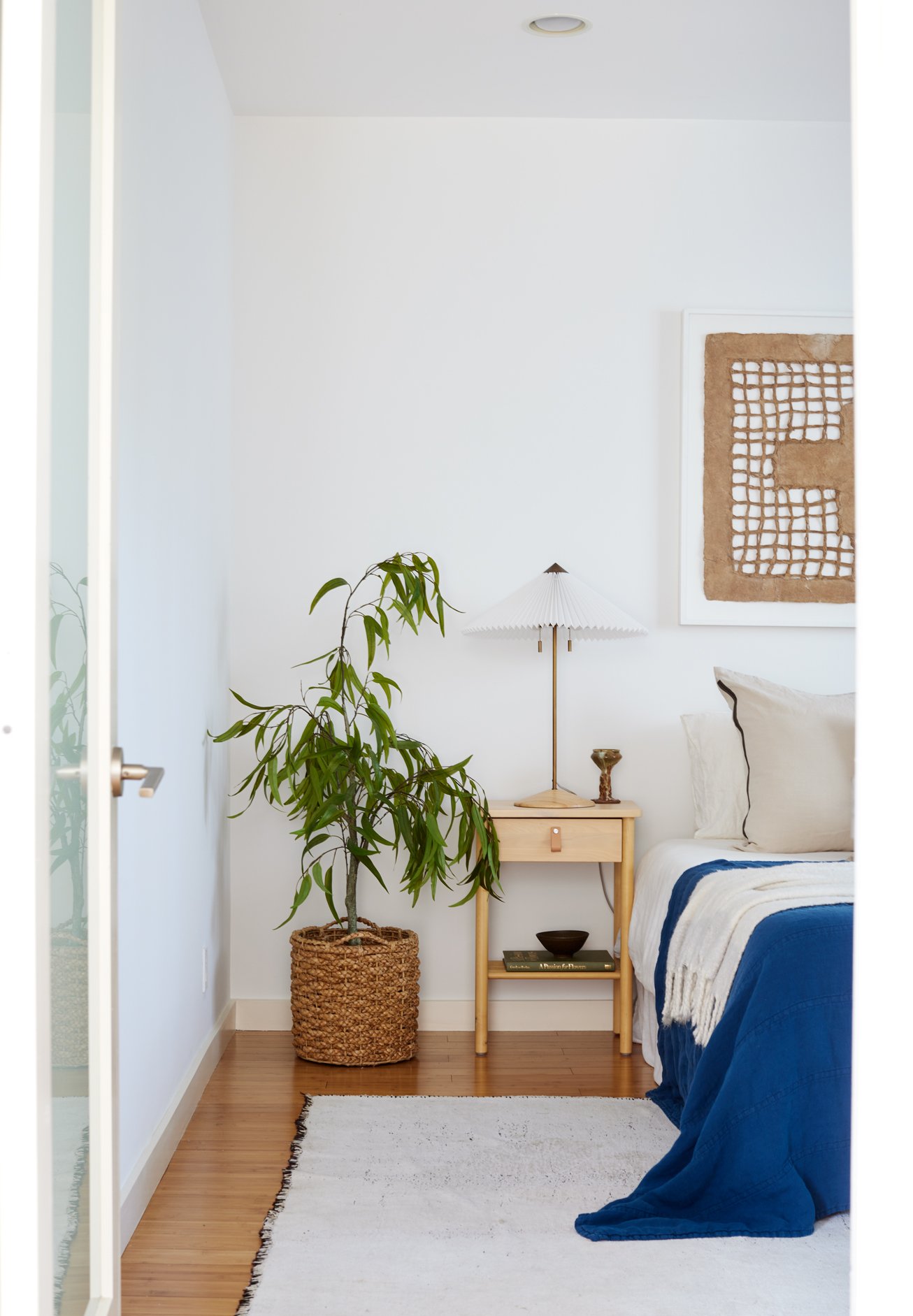 A cozy bedroom corner with a bed, floor lamp, wooden nightstand, large potted plant in a woven basket, framed modern artwork, and a white rug on hardwood flooring.