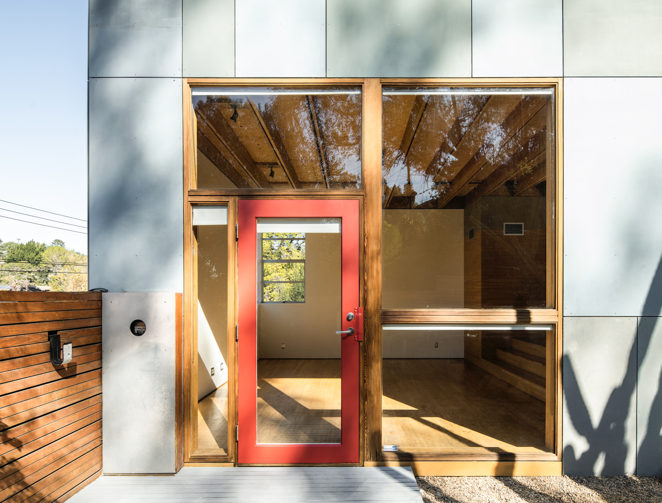 Modern building entrance with red door, large glass windows, and wooden accents, surrounded by trees and clear sky.