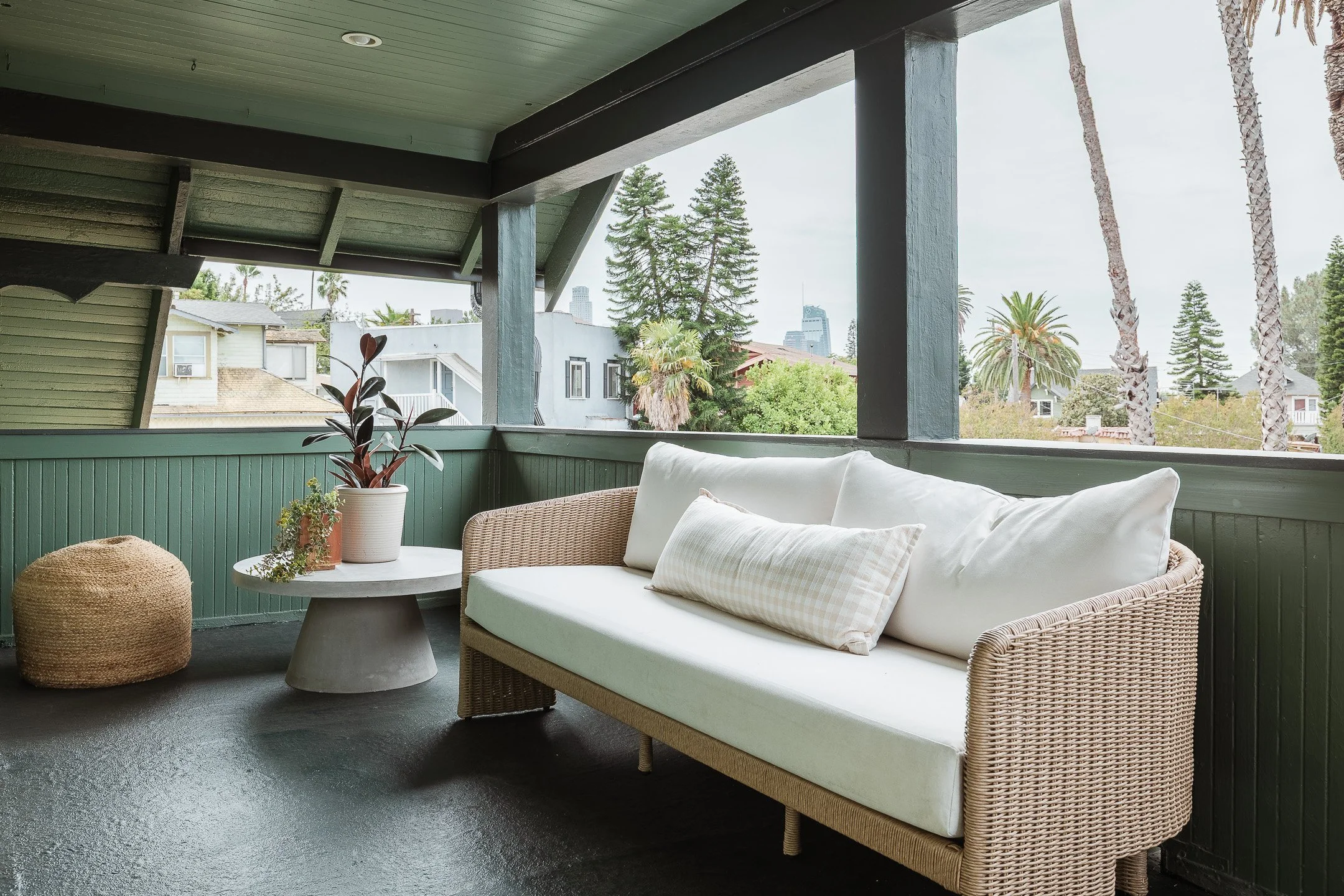 Outdoor balcony with green wooden walls, a beige wicker sofa with white cushions and a long pillow, a small round gray concrete table with potted plants, a woven pouf, and a view of lush trees and buildings in the distance.