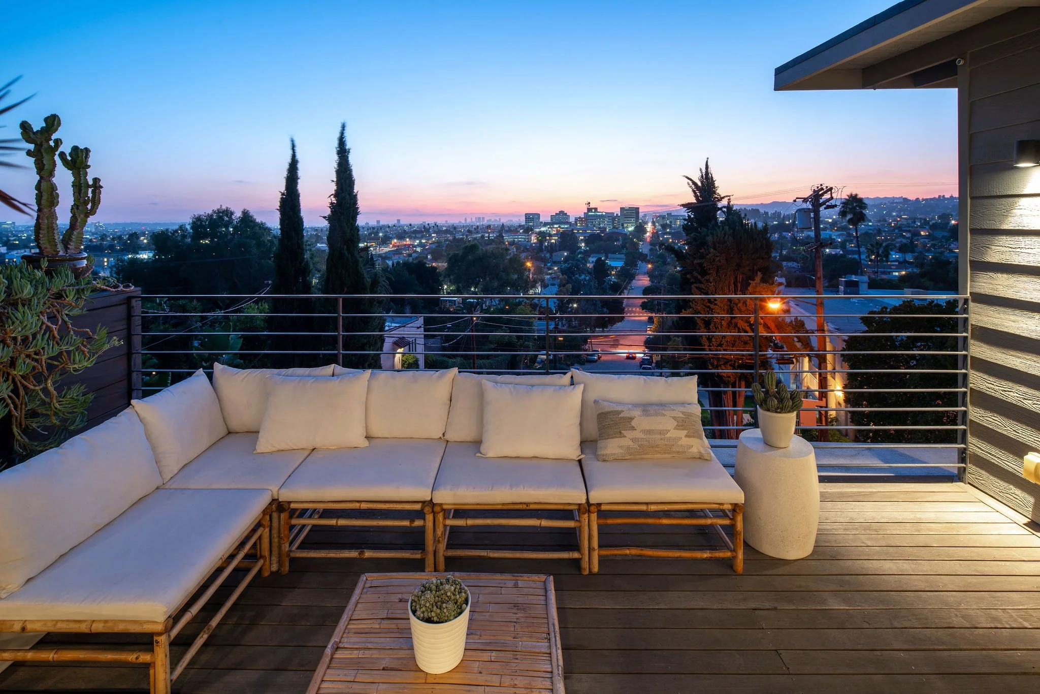 Outdoor rooftop patio with a sectional sofa, potted plants, and a city view at sunset.