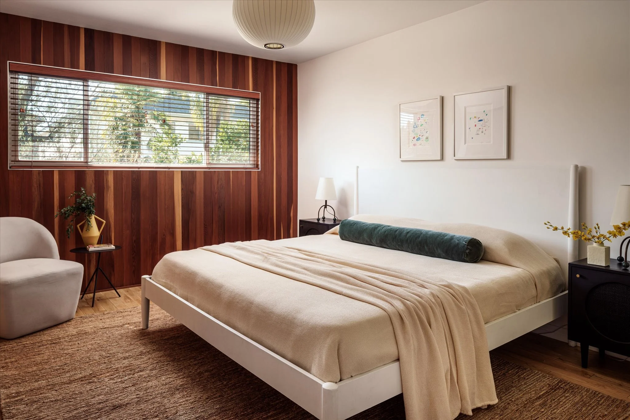 Modern bedroom with a bed, beige bedding, green bolster, white walls, artwork, black nightstands, lamps, a beige armchair, a small side table with a plant, and a wooden accent wall with window blinds.