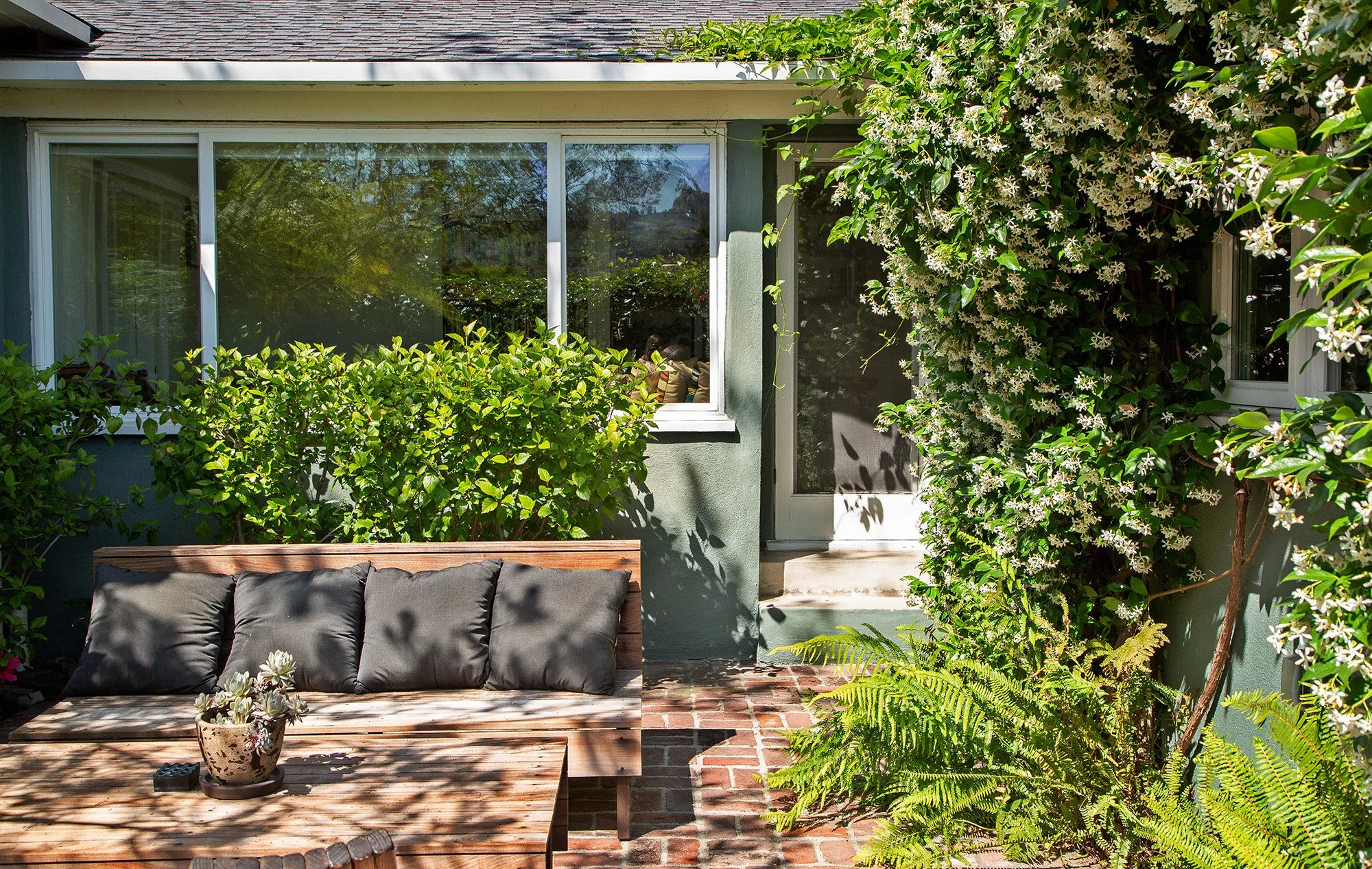 A cozy outdoor patio with a wooden bench, lush greenery including bushes and a flowering vine on the right, a potted succulent on a wooden table, outside a house with large windows and a door, surrounded by natural sunlight and shadow patterns.