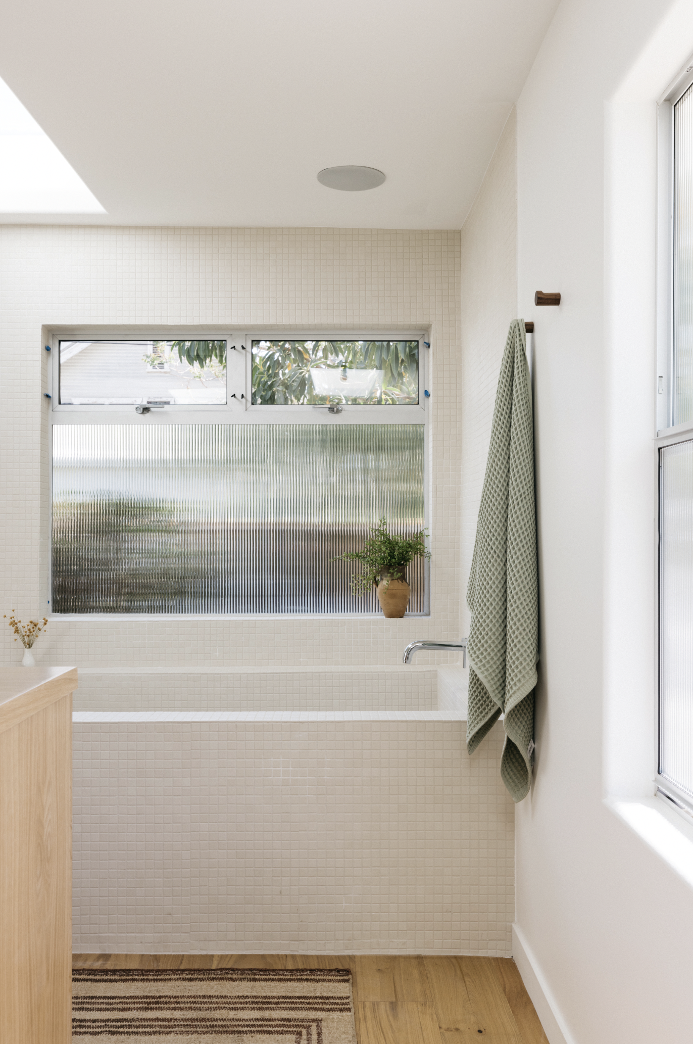 A bathroom with a small tub beneath a large frosted window, a potted plant on the window sill, and two green towels hanging on a wall hook.