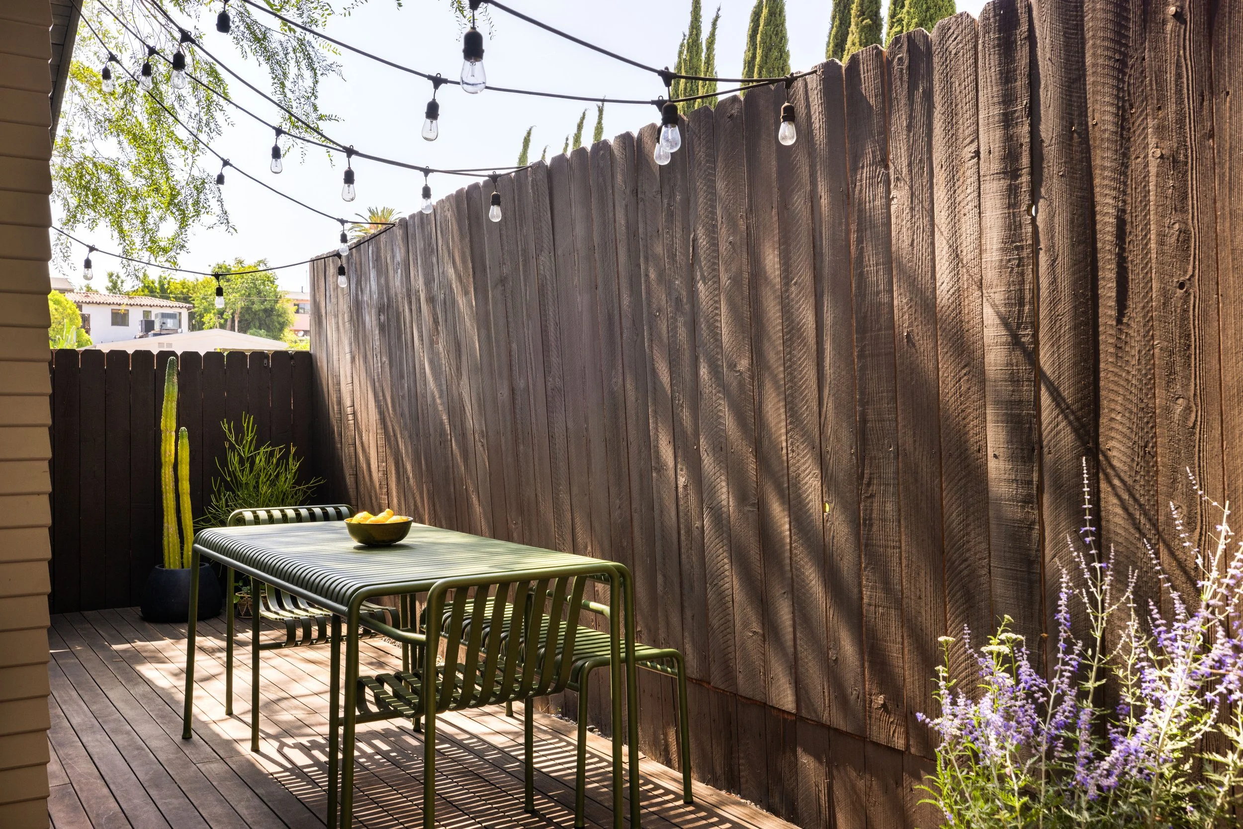 A small outdoor patio with wooden decking, a table with a bowl of lemons, metal chairs, potted cactus plant, purple flowering plants, and string lights hanging above, enclosed by wooden fences.
