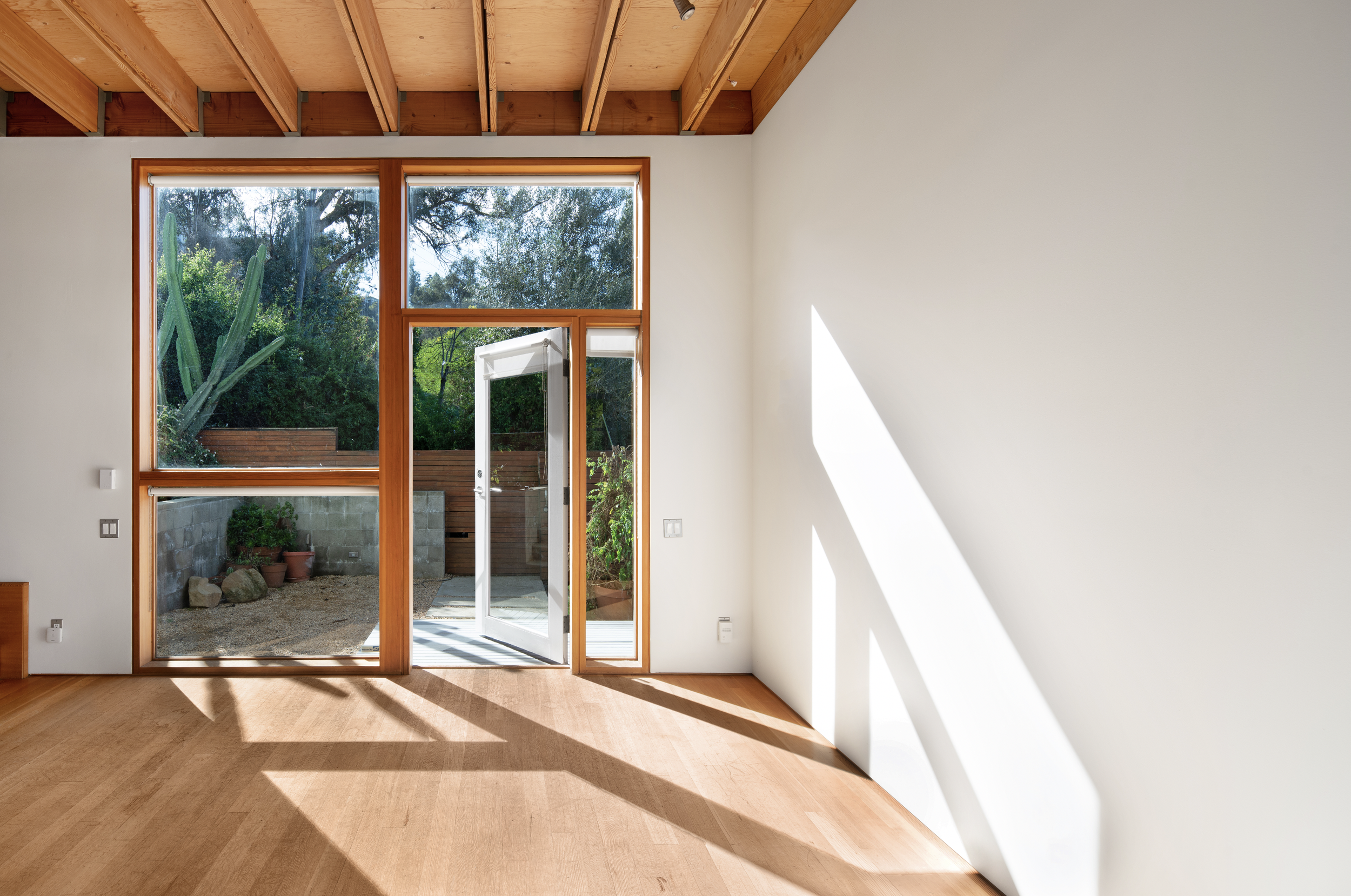 Empty room with large glass doors and windows showing an outdoor patio with plants, rocks, and a wooden fence; sunlight casting shadows on the wooden floor; ceiling with exposed wooden beams.