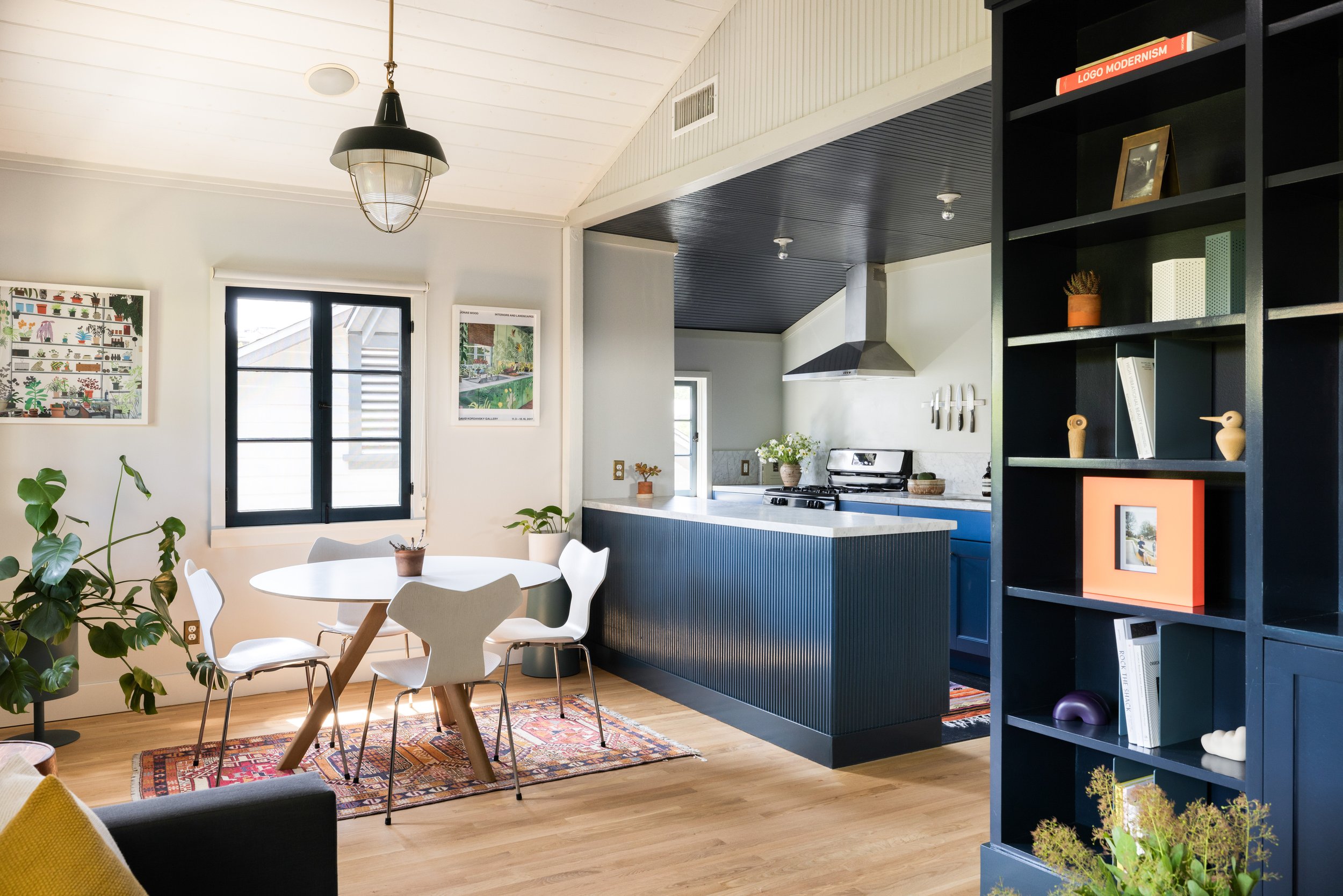 Open-concept living room and kitchen with white walls, wooden floors, black shelving unit, dining table with four chairs, plants, and modern decor.
