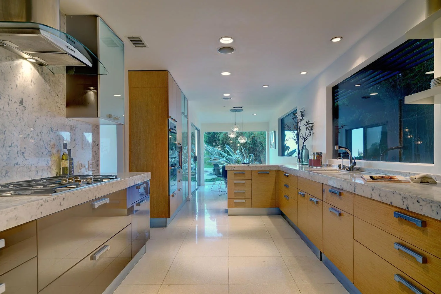 Modern kitchen with wooden cabinets, marble countertops, and large windows looking into a backyard with lush greenery