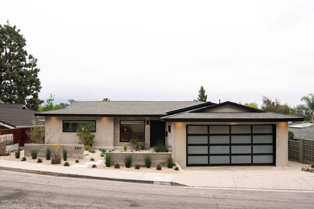Modern single-story house with a flat roof, large front window, and attached garage with a frosted glass garage door, surrounded by desert-style landscaping and small plants.