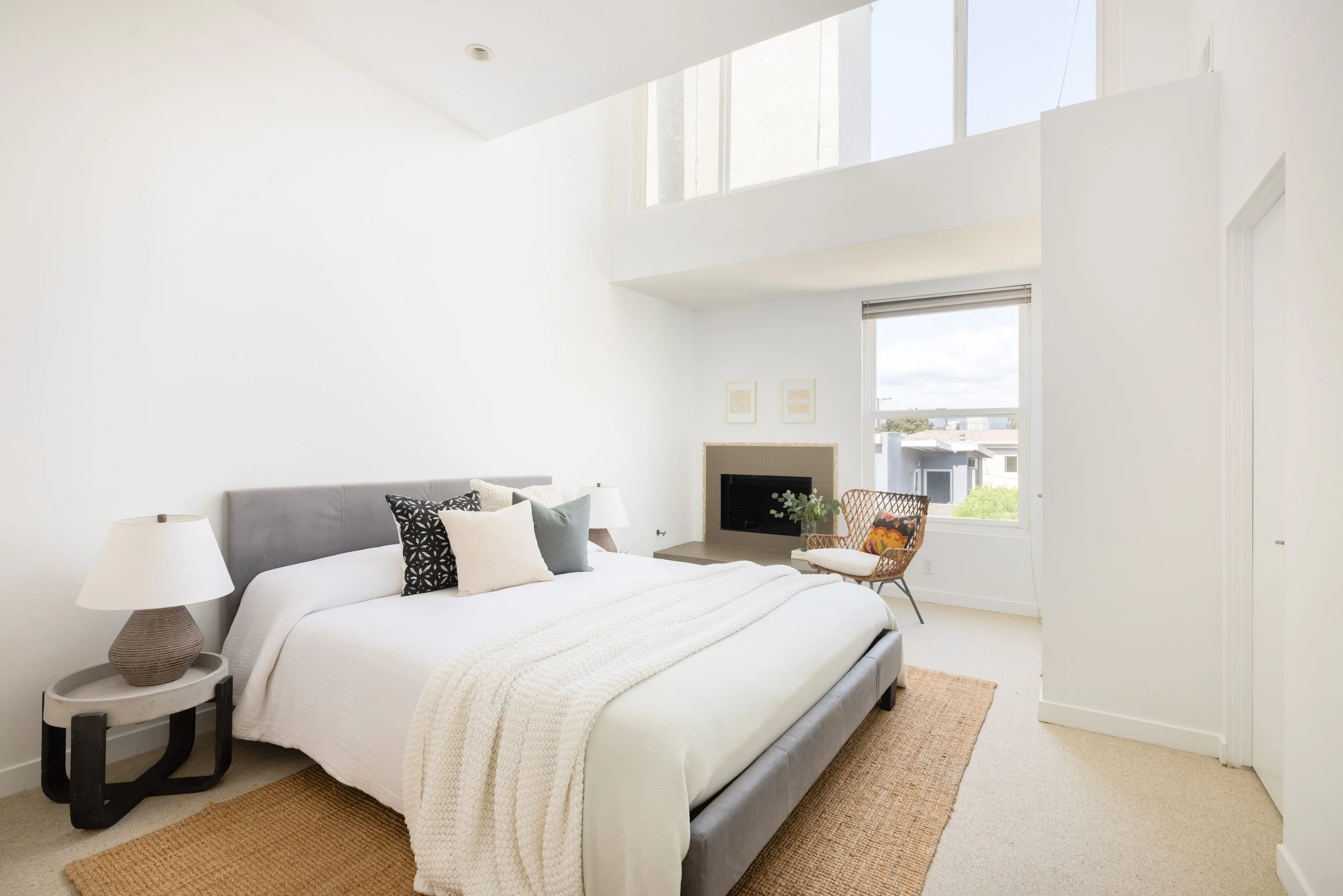 Bright, modern bedroom with white walls, large windows, gray bed with white bedding and decorative pillows, side table with lamp, wicker chair near fireplace, and simple decor.