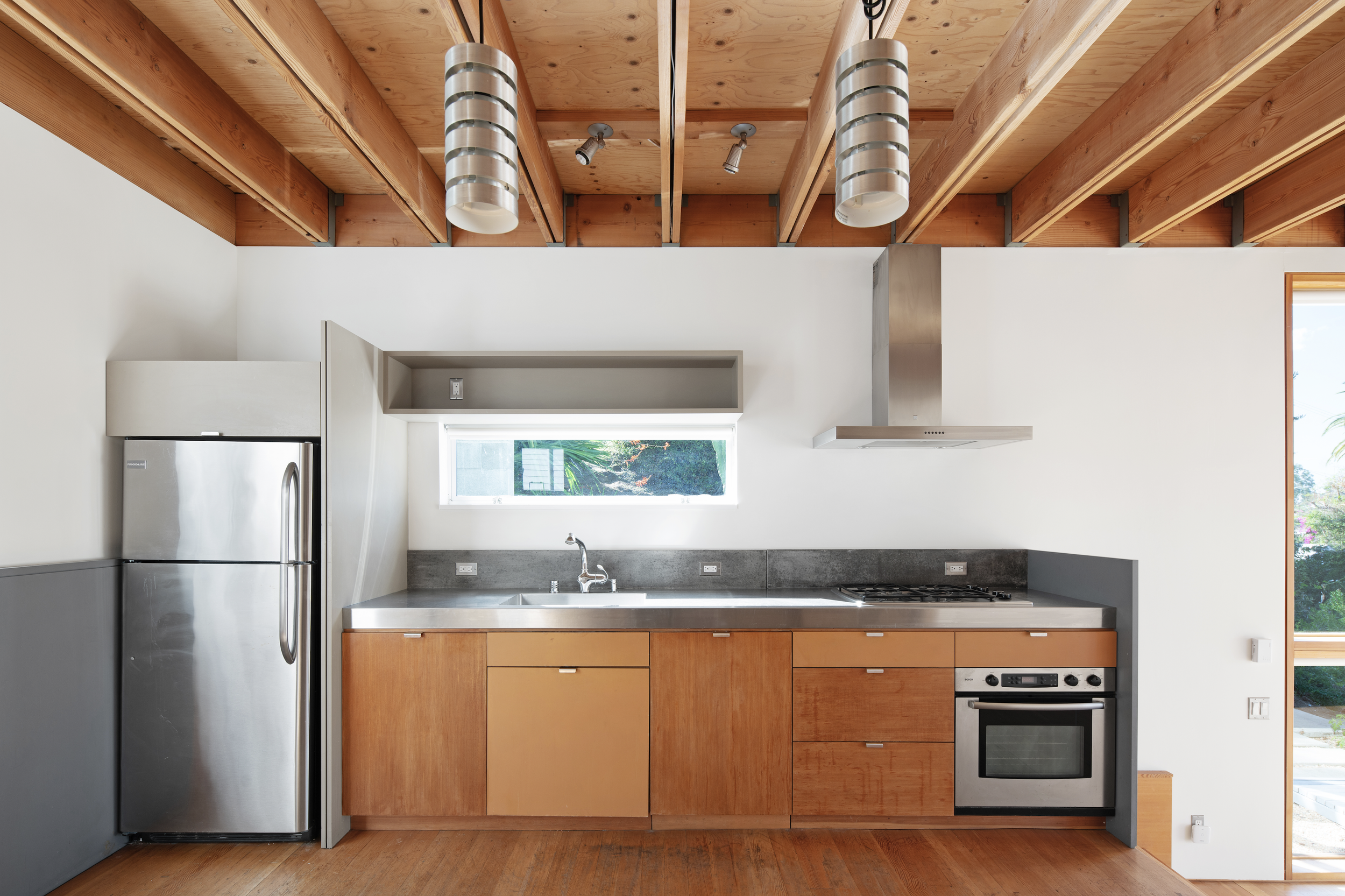 Minimalist kitchen with stainless steel refrigerator, wooden cabinets, gray countertop, small window above the sink, and stainless steel range hood, with exposed wooden beam ceiling.