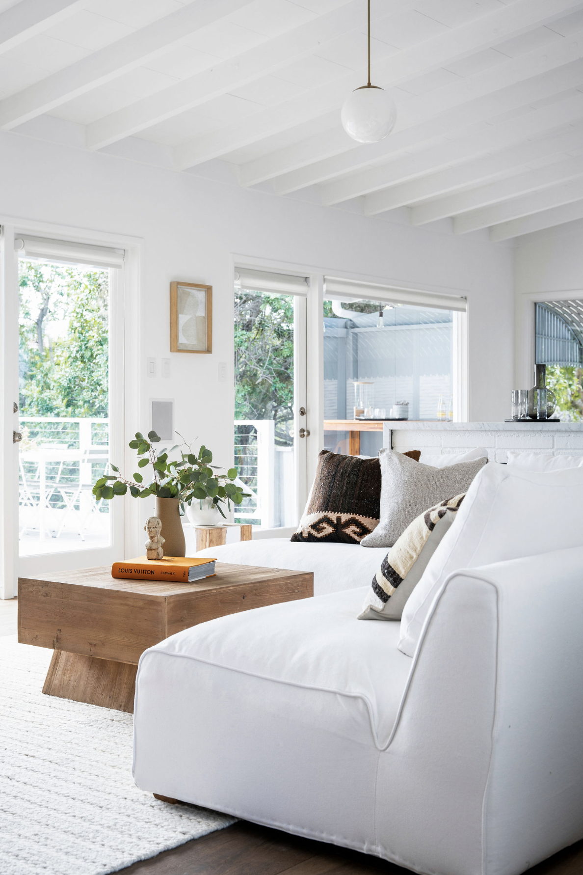 Bright living room with white sofa, wooden coffee table, and large windows showing outdoor trees and patio.