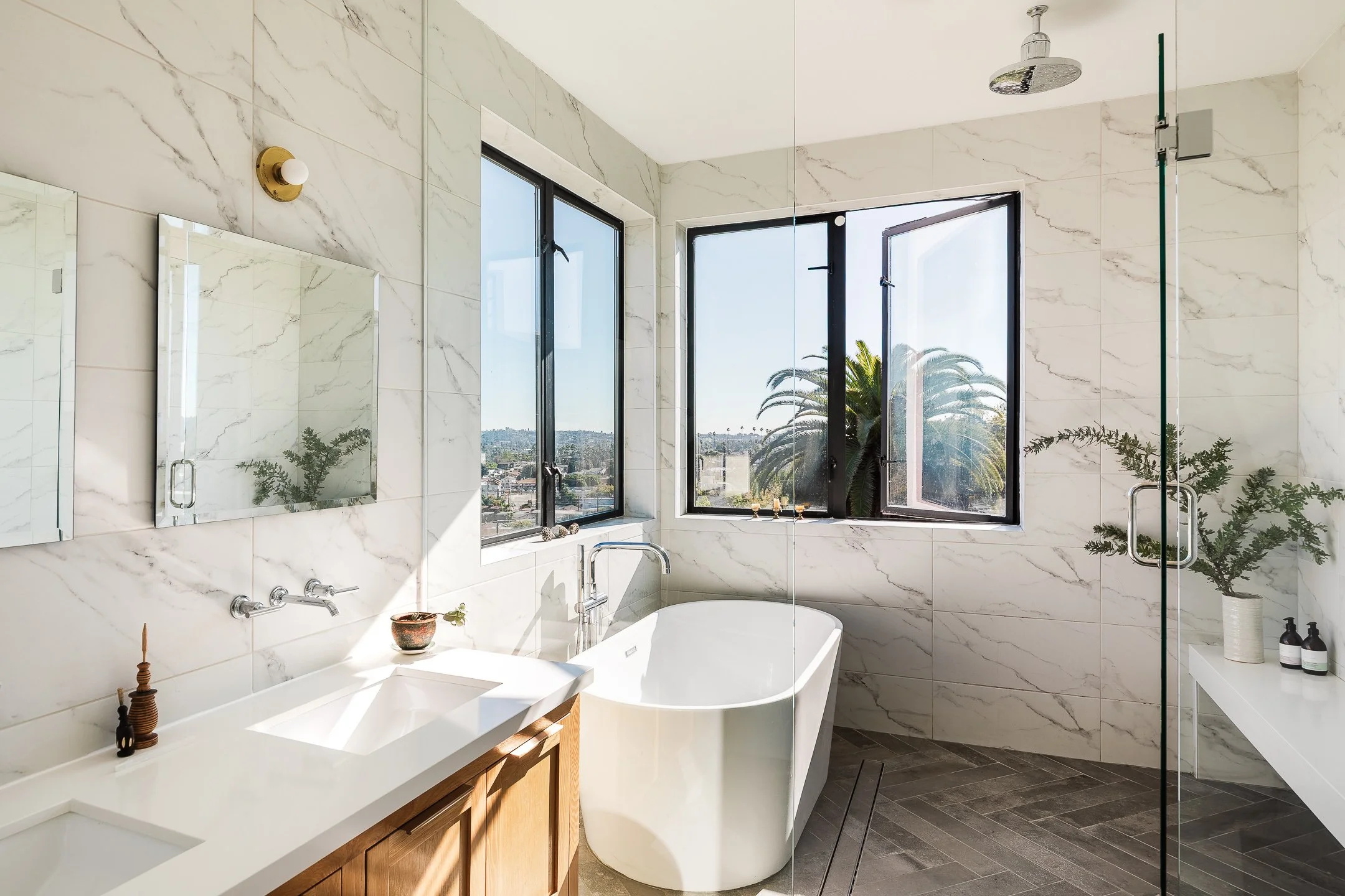 Modern bathroom with white marble walls, a free-standing bathtub, a double sink vanity with a wooden cabinet, large windows overlooking palm trees, and a glass shower enclosure.