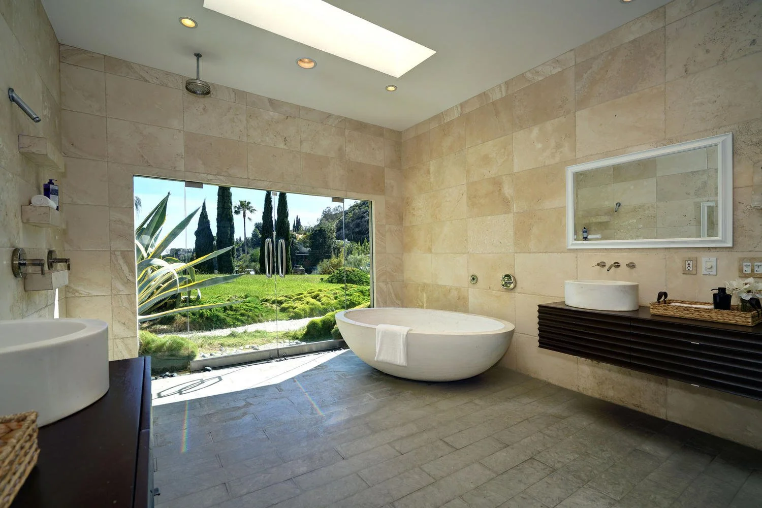 Modern bathroom with large glass sliding doors open to lush green outdoor garden, freestanding oval bathtub, wall-mounted sink with a mirror, beige tiled walls, and tiled floor, natural light streaming in.