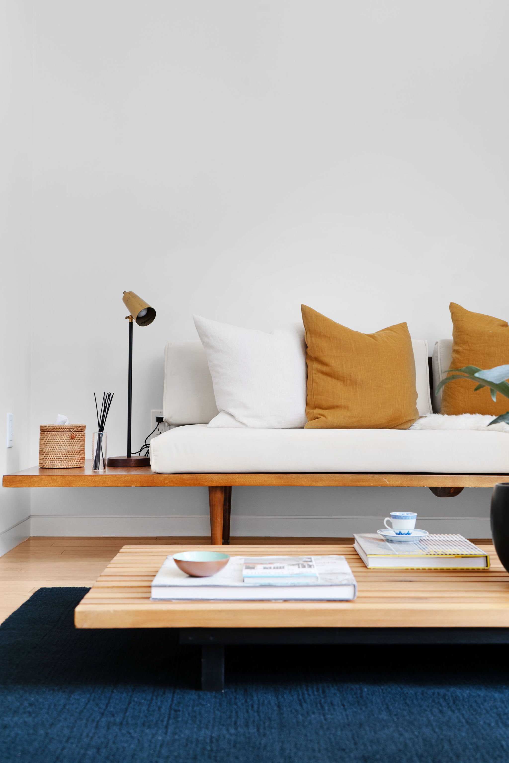 Living room with a white sofa, mustard pillows, a wooden coffee table with magazines and cups, a black rug, lamp, tissue box, and reed diffuser on a side table against a white wall.