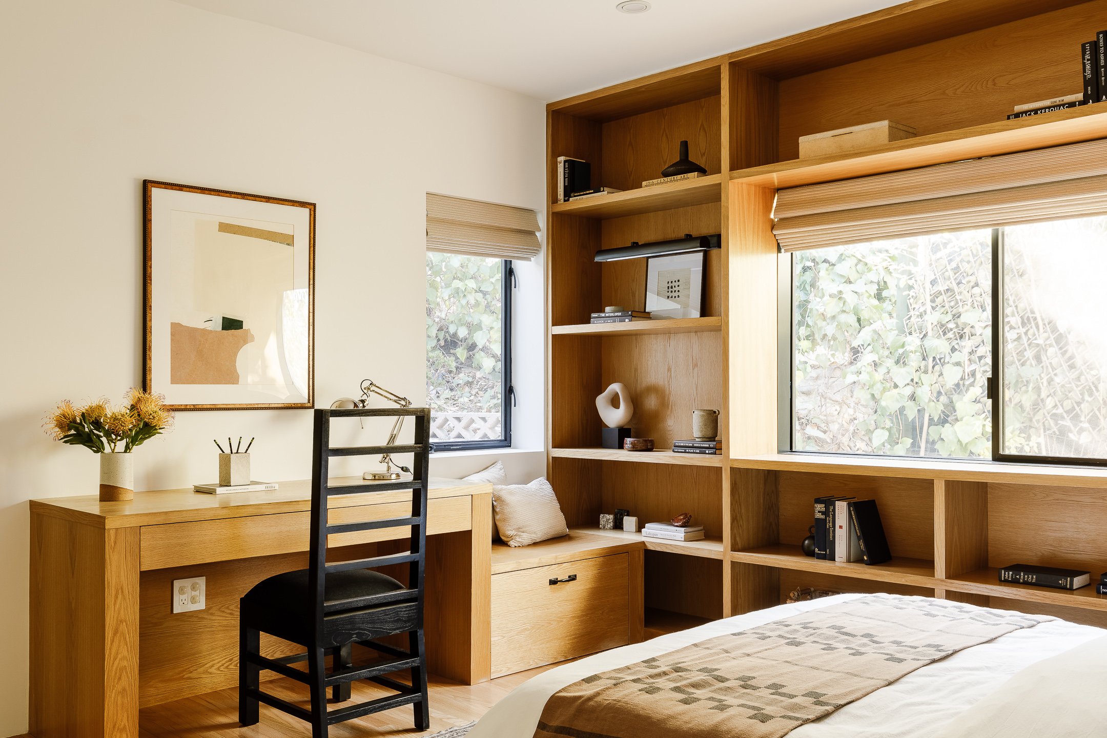 A bedroom with a wooden desk and chair, a built-in wooden bookcase, a window with blinds, a bed with a beige blanket, and decorative items like plants and sculptures.