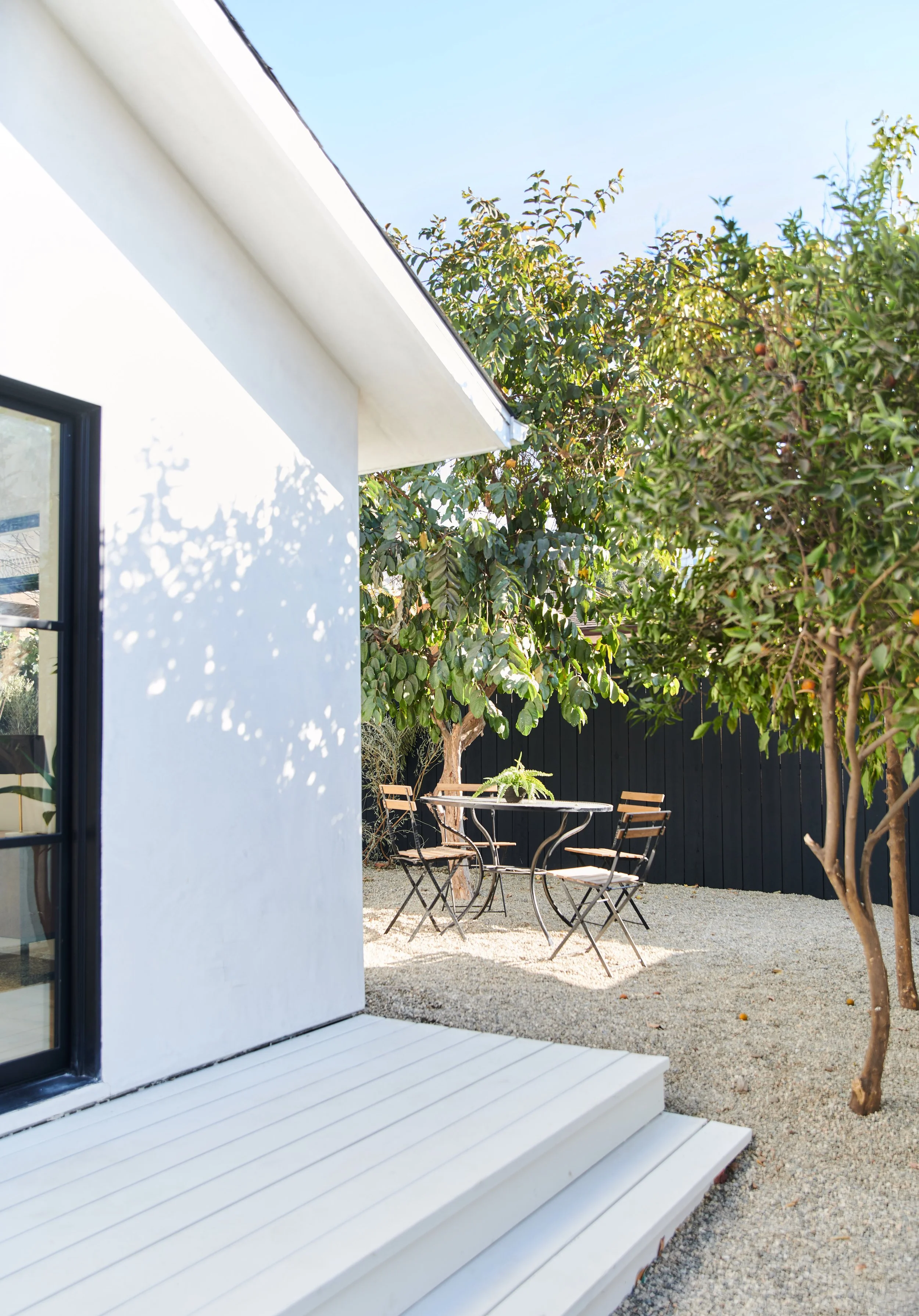 A white house with a small porch and black-framed window. Outside, there is a gravel yard with a round metal table and four wooden chairs, surrounded by green trees and a black fence.