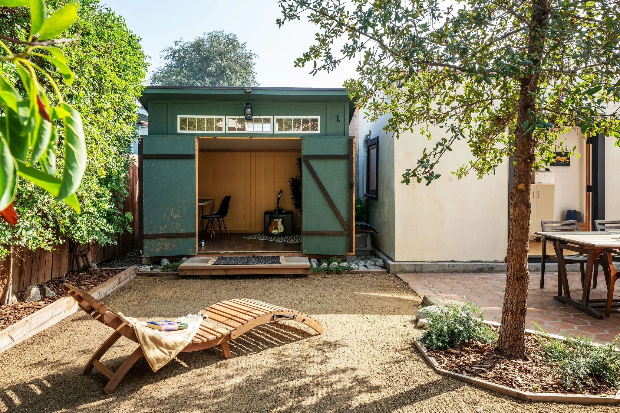 Backyard with a small green shed, wooden deck, lounge chair with towel, mature trees, a white house with porch, outdoor table and chairs, and landscaping with plants and mulch.