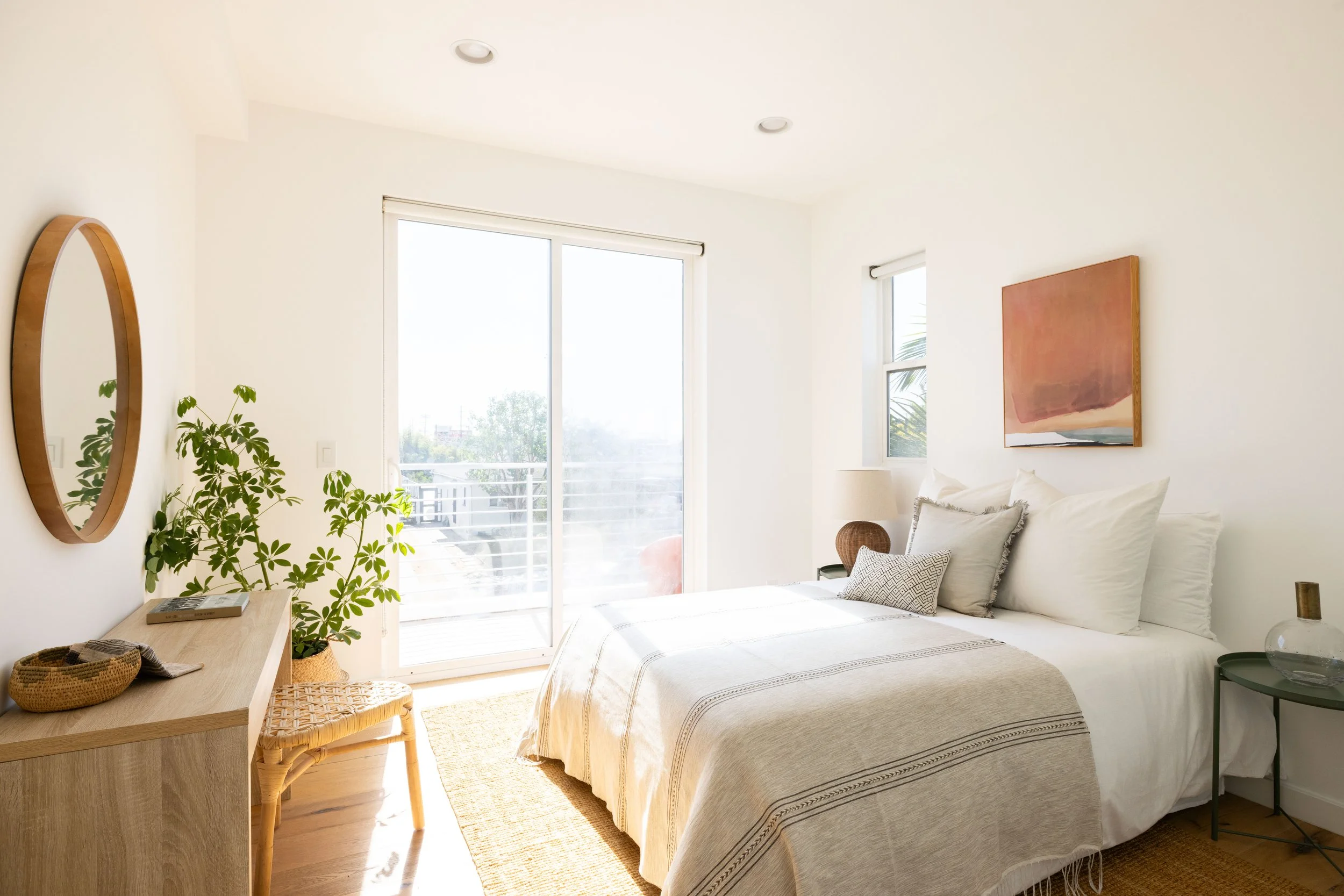 Bright bedroom with large sliding glass door, white walls, and minimal decor, including a bed with white bedding, a side table with a lamp, and artwork on the wall.