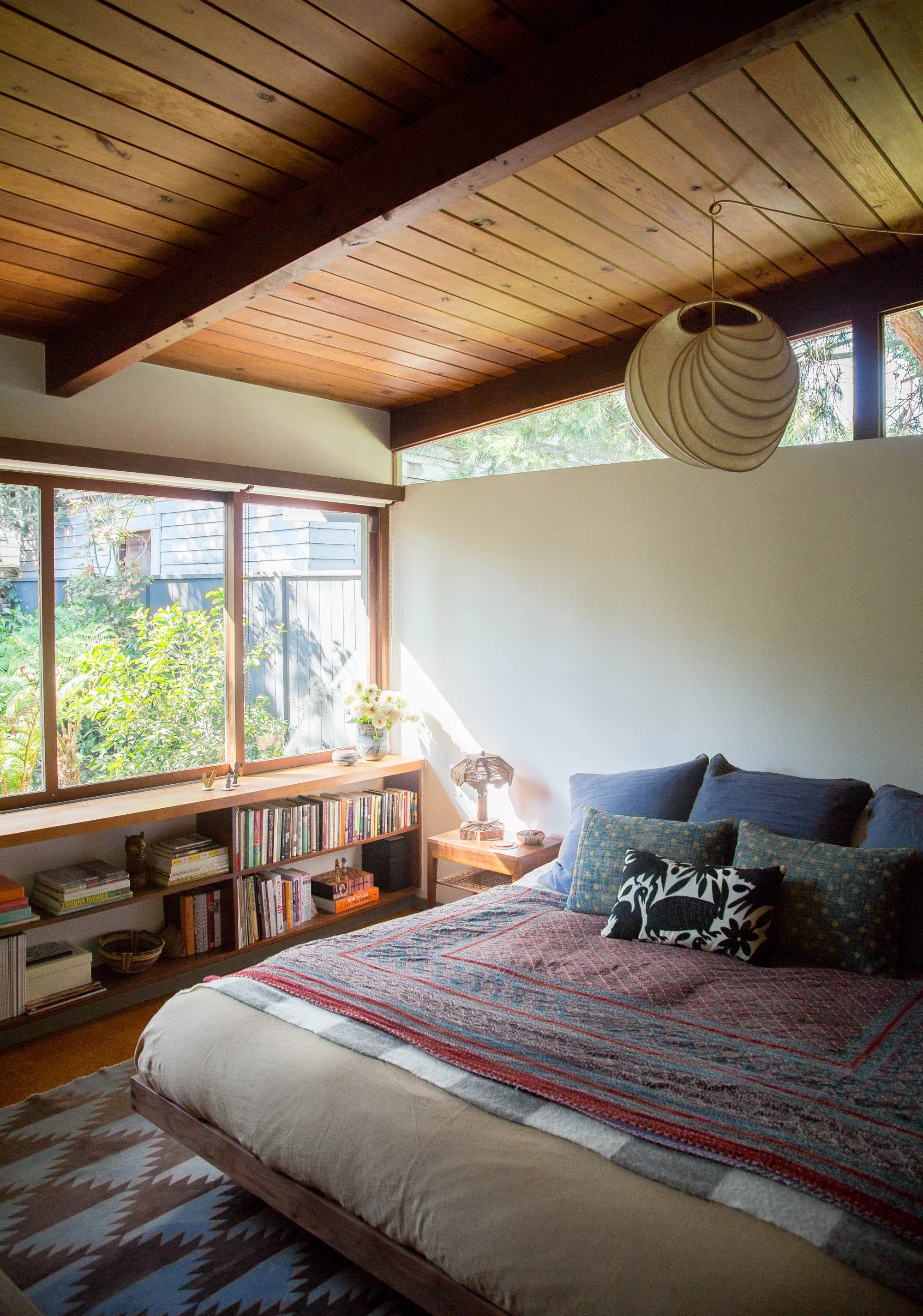 Cozy bedroom with wooden ceiling, large window with greenery outside, bookshelf, side table with lamp, and bed with patterned pillows.